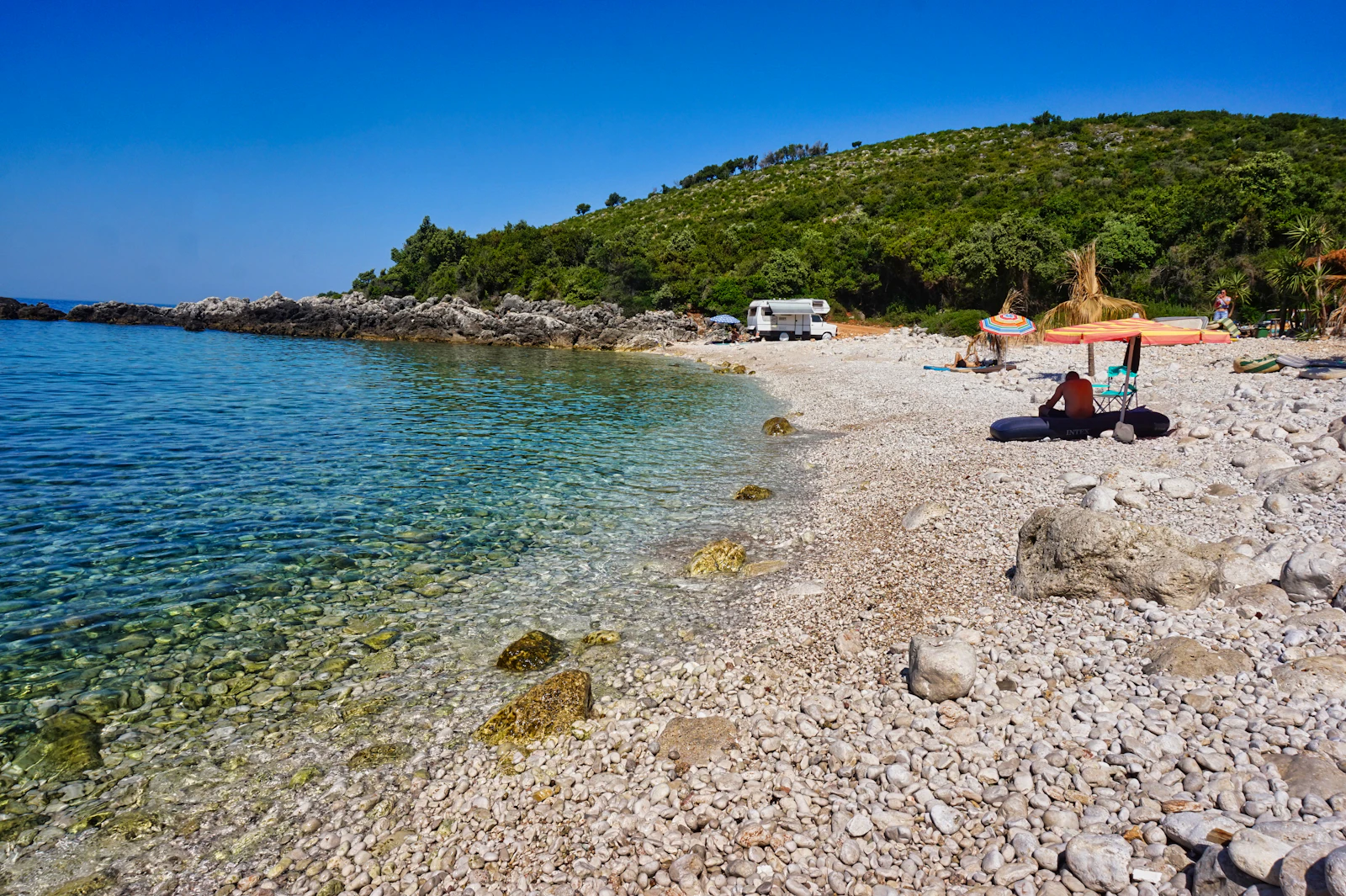 A pebble beach on the Himarë coast, clear water and the green hills behind