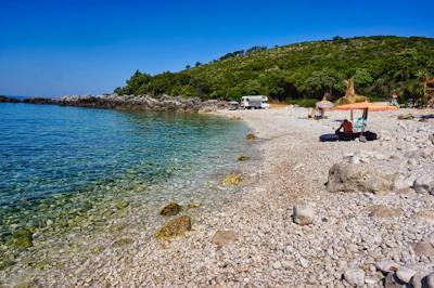 A pebble beach on the Himarë coast, clear water and the green hills behind