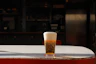 A single pint glass of lager with a thick foam head on a speckled bar table, lit by low evening sun