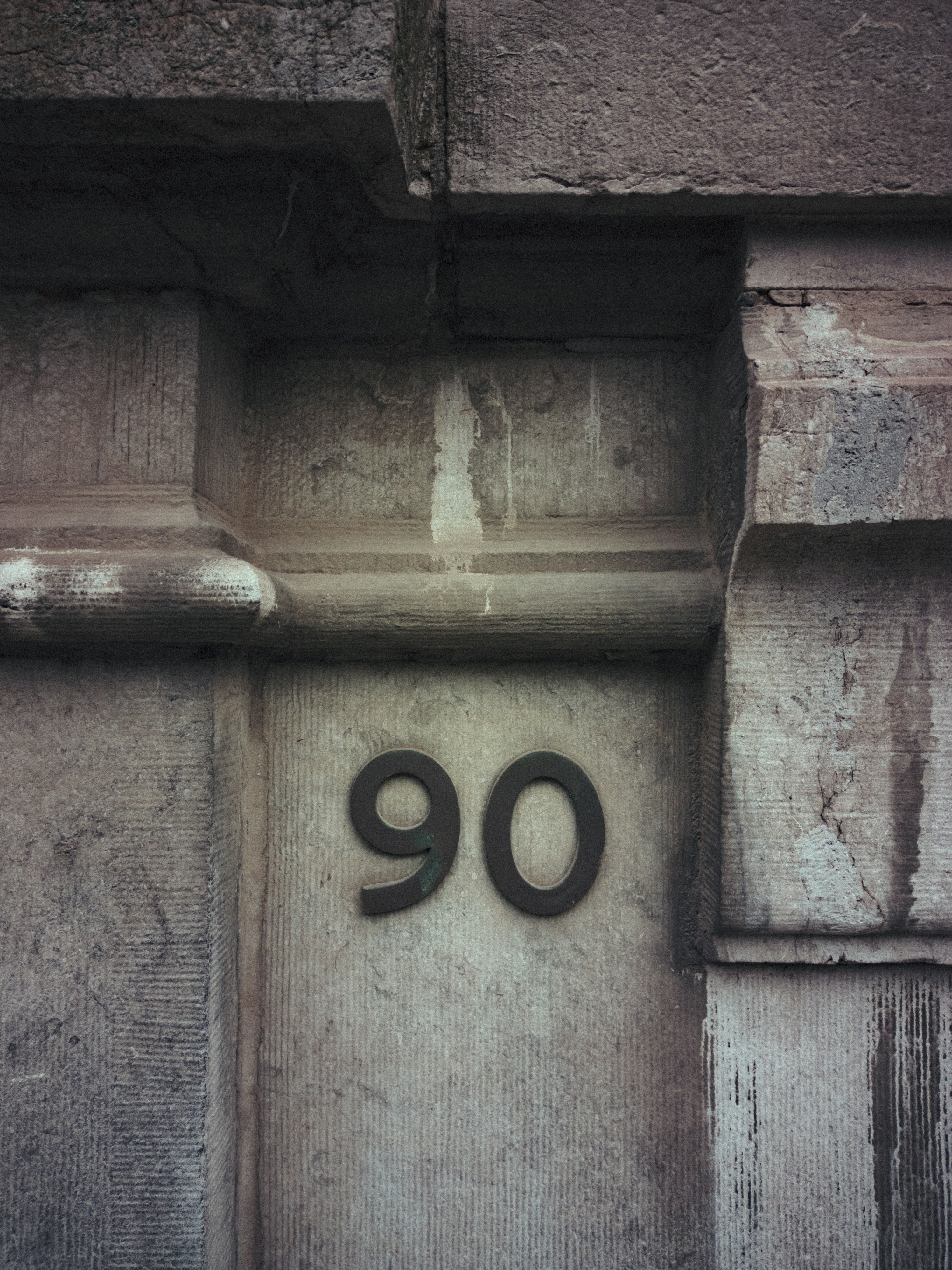 House number ninety (90) on a textured gray surface with dirt stains visible. The numbers are made of a metal base, painted black and the metal is oxidized