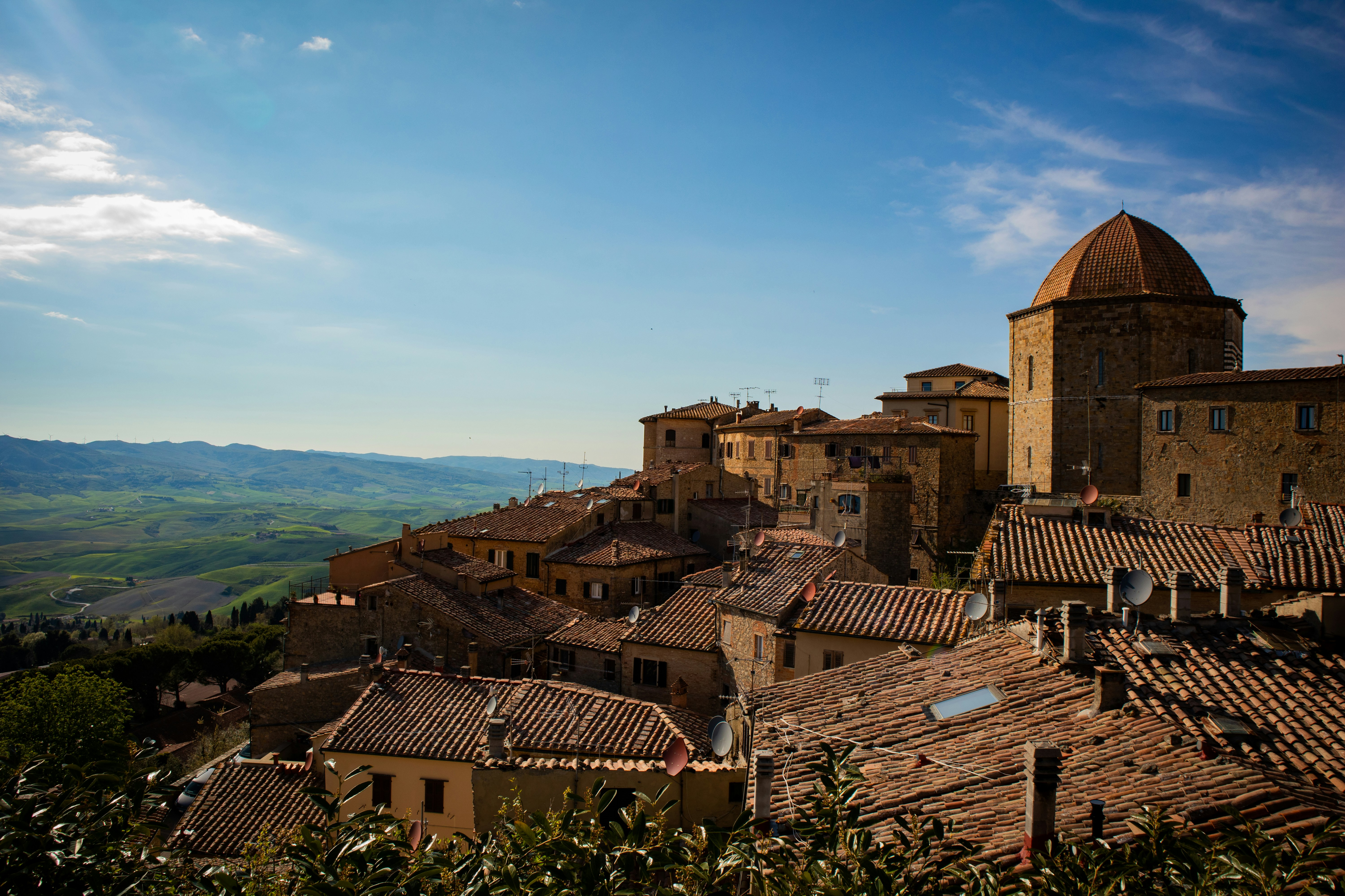 The image portrays the charming hilltop town of Volterra, Tuscany, Italy. It features terracotta rooftops, stone buildings, and the prominent dome of a historic structure. The town is nestled amidst rolling green hills, with a clear blue sky and scattered clouds enhancing the picturesque atmosphere. The lighting highlights the warm tones of the architecture and the vibrant greenery, creating a serene and timeless scene that reflects the rich medieval heritage of Volterra.