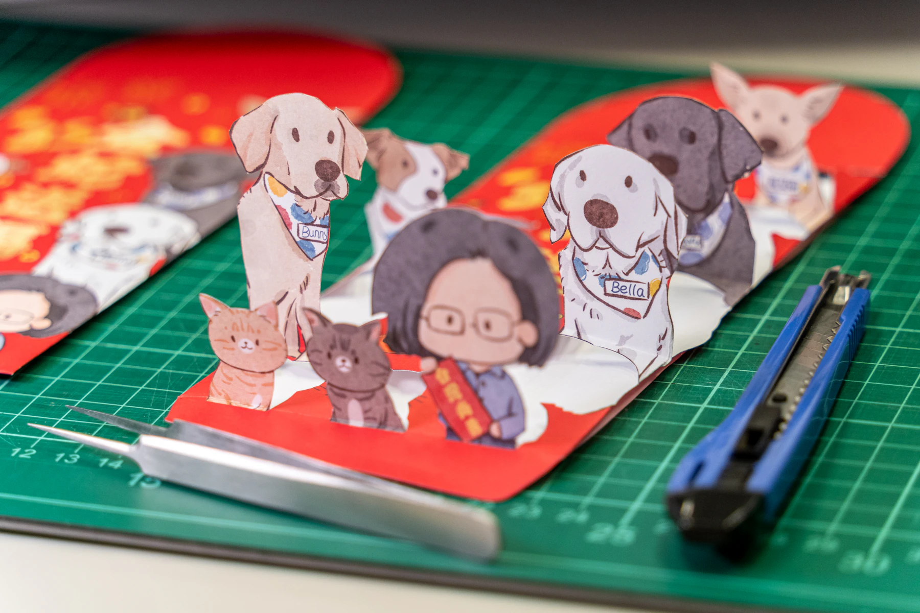 Printed paper figures of pets and a person, cut out and standing on a green cutting mat beside a knife and tweezers