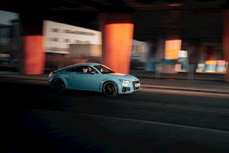 A blue car driving down a street next to tall buildings