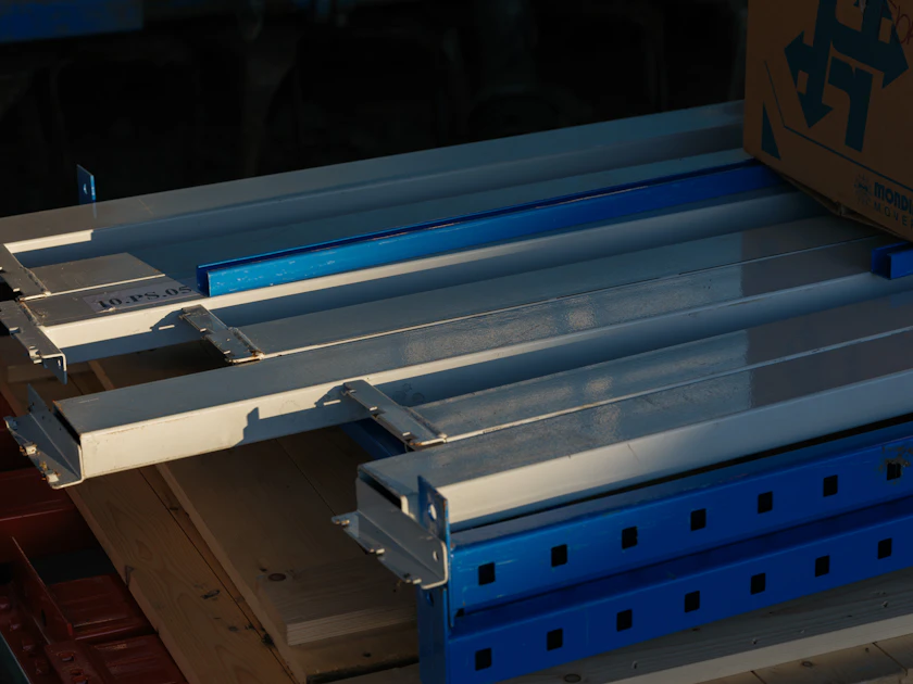 Lengths of metal profile laid out on a wooden pallet beside blue steel racking beams.