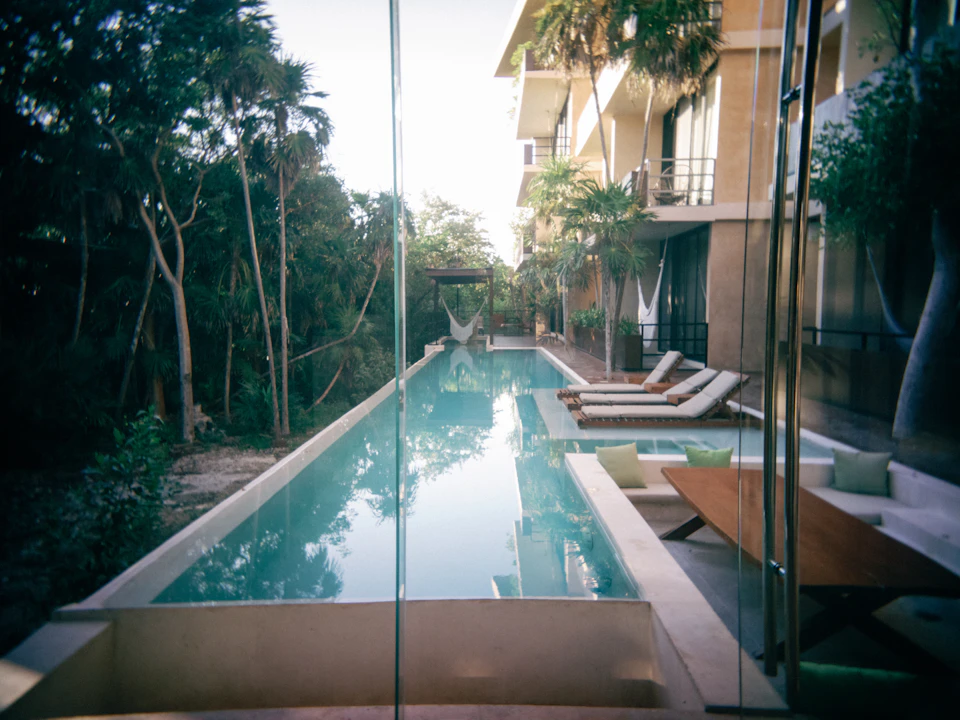 The image depicts a luxurious outdoor pool area with a large, rectangular pool filled with clear blue water and surrounded by white lounge chairs arranged on both sides. The pool is situated within an apartment complex, with palm trees visible in the background. A balcony can be seen above the pool, offering a view of the surrounding greenery.
