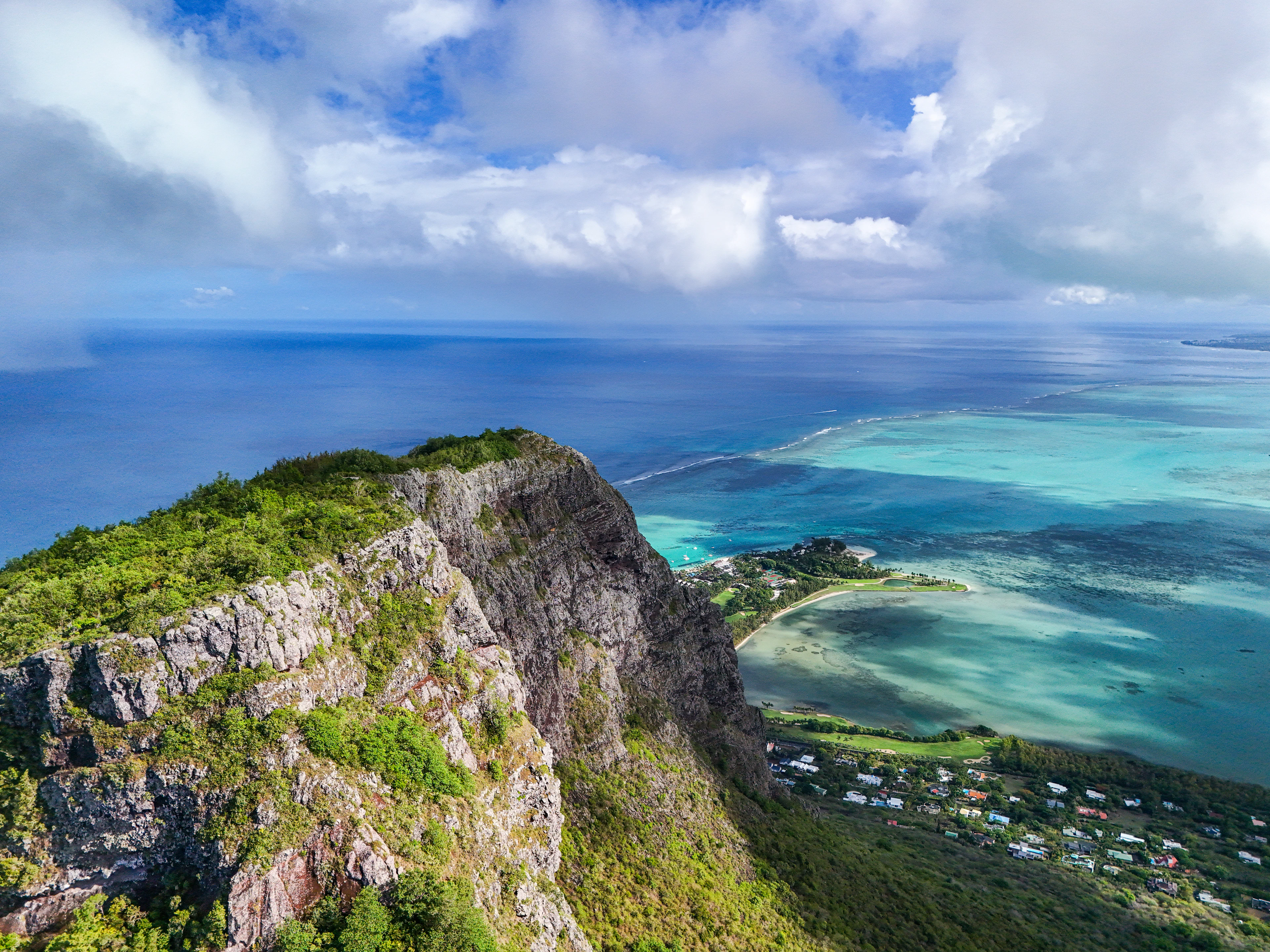 A dramatic aerial from a cliff edge looks down over a turquoise lagoon and coastal village.
