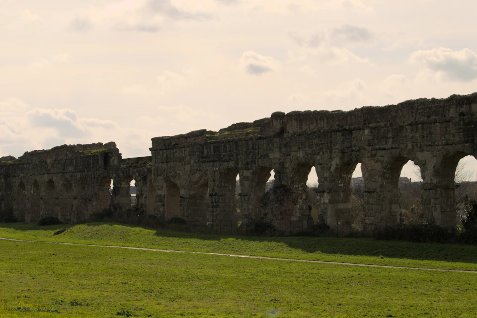 A long line of ancient Roman aqueduct arches in soft winter light, standing over a wide green lawn crossed by a footpath
