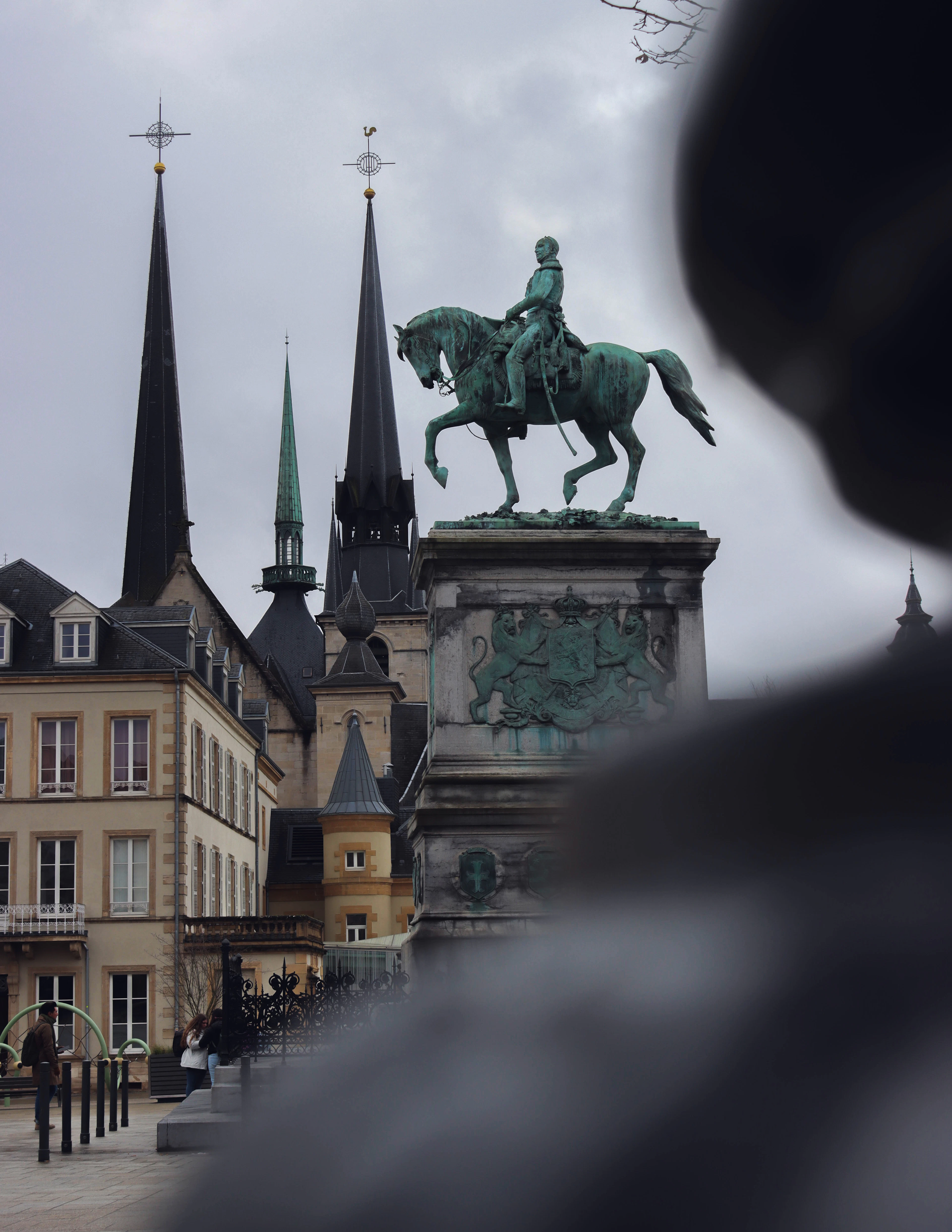 A bronze equestrian statue on a plinth stands with the city's church spires rising behind it under a grey sky.