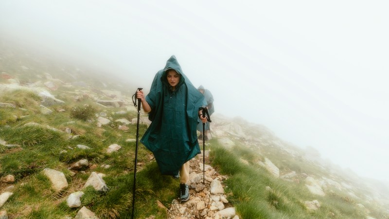 Hiker in a long dark green hooded poncho walking up a foggy grassy mountain path with trekking poles