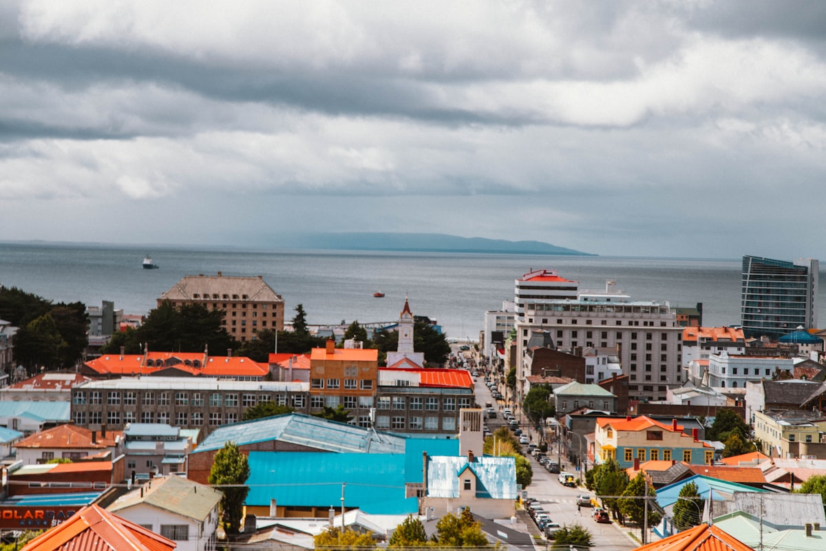 A southern port city seen from a hillside, its red, blue and green roofs running down to a wide grey channel with a ship on it and low hills on the far shore under heavy cloud