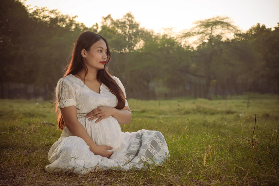 A pregnant woman in a white dress sitting in a grassy field at golden hour
