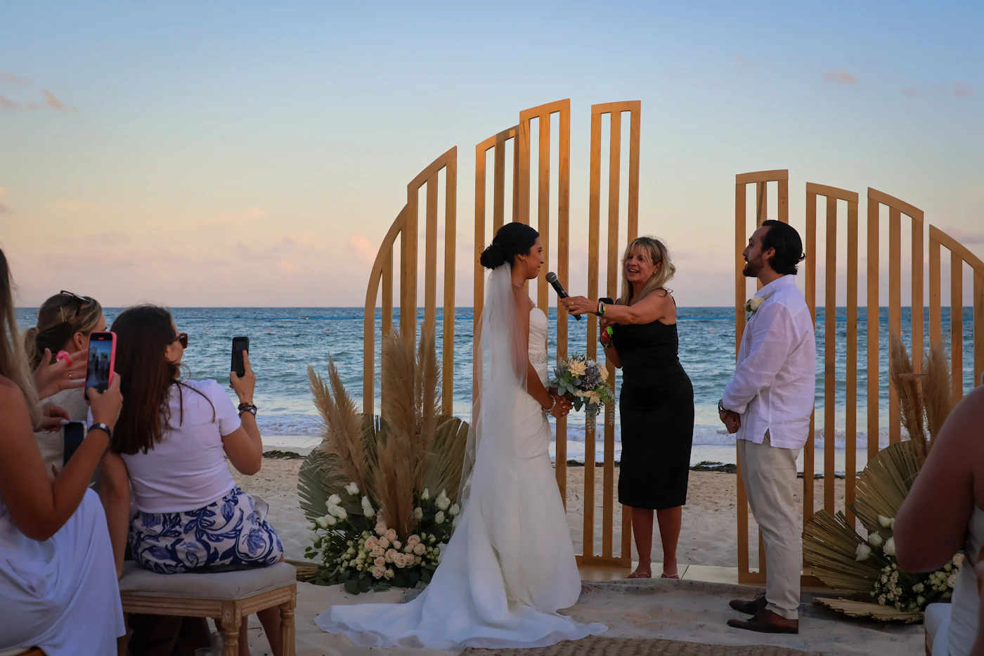 A bride and groom exchanging vows at a Riviera Maya beach wedding ceremony at dusk — a wooden sculptural arch with pampas grass and white flowers, guests seated on either side, the Caribbean sea fading from turquoise to blue behind them