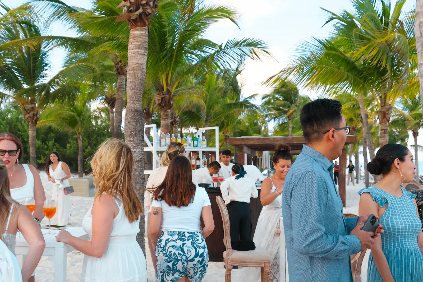 Guests in summer attire mingling with drinks at a beachside reception under palm trees