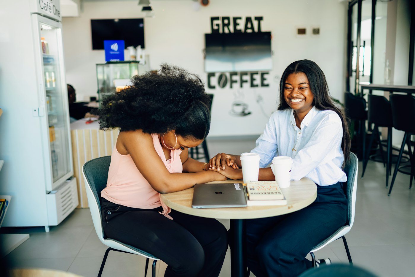 Duas mulheres conversam numa mesa de café, com o notebook aberto entre elas.