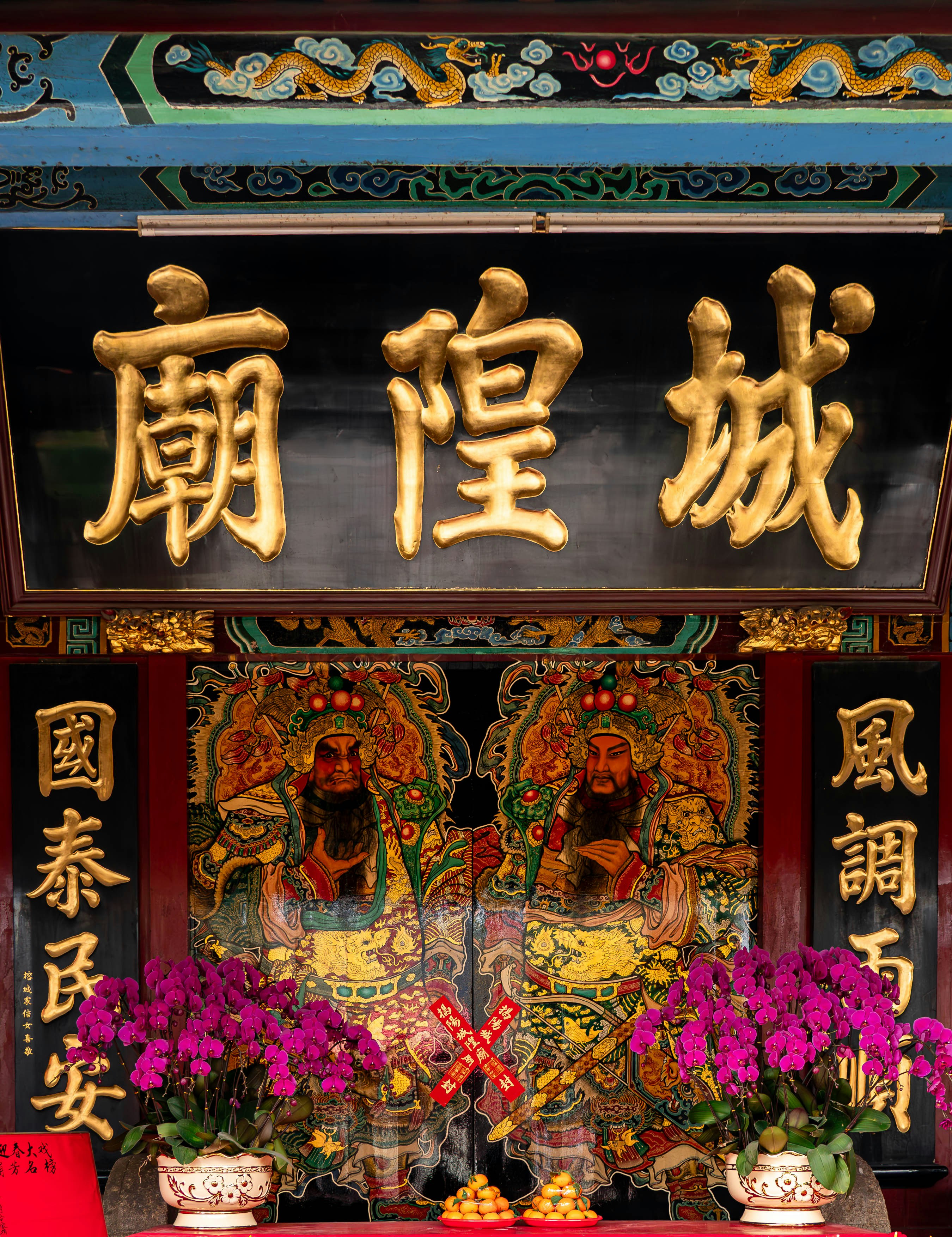 An ornate entrance to a traditional Chinese temple features large, gilded Chinese characters (廟隍城 - Chenghuang Temple/City God Temple) on a plaque above intricately painted doors depicting fierce door gods. Flanking the doors are vertical plaques with golden calligraphy expressing wishes for peace and favorable conditions (國泰民安, 風調雨順). Offerings of vibrant magenta orchids and small oranges rest on an altar table in front, completing this colorful and detailed display of traditional religious art and symbolism.