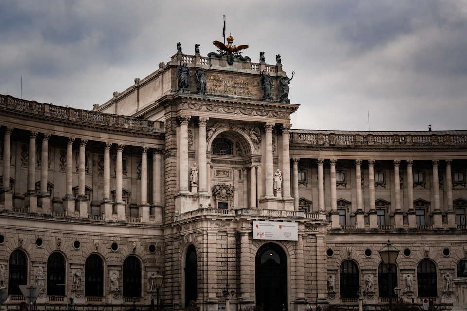 The image shows the majestic Schönbrunn Palace, a renowned landmark located in Vienna, Austria. The palace is an architectural marvel with its grandeur and intricate design. It features numerous columns that add to its imposing presence. A large golden eagle statue stands atop the palace, symbolizing power and authority. The perspective of the photo is from street level, looking up at the palace, emphasizing its height and dominance in the cityscape.

The sky above the palace is filled with clouds, casting a soft light over the scene. The image code 'sa_1296' may be a reference to the specific angle or viewpoint from which the photograph was taken.