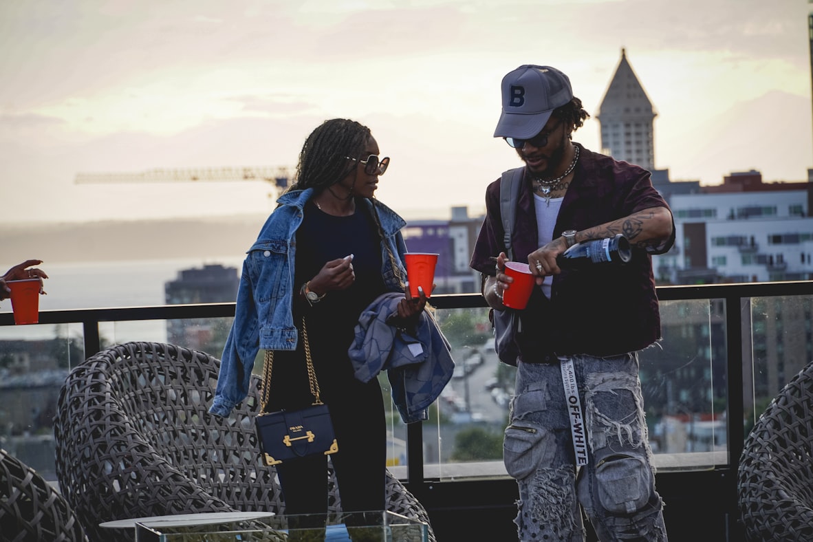 A pair of travelers standing close together on a rooftop at dusk, the city skyline behind them
