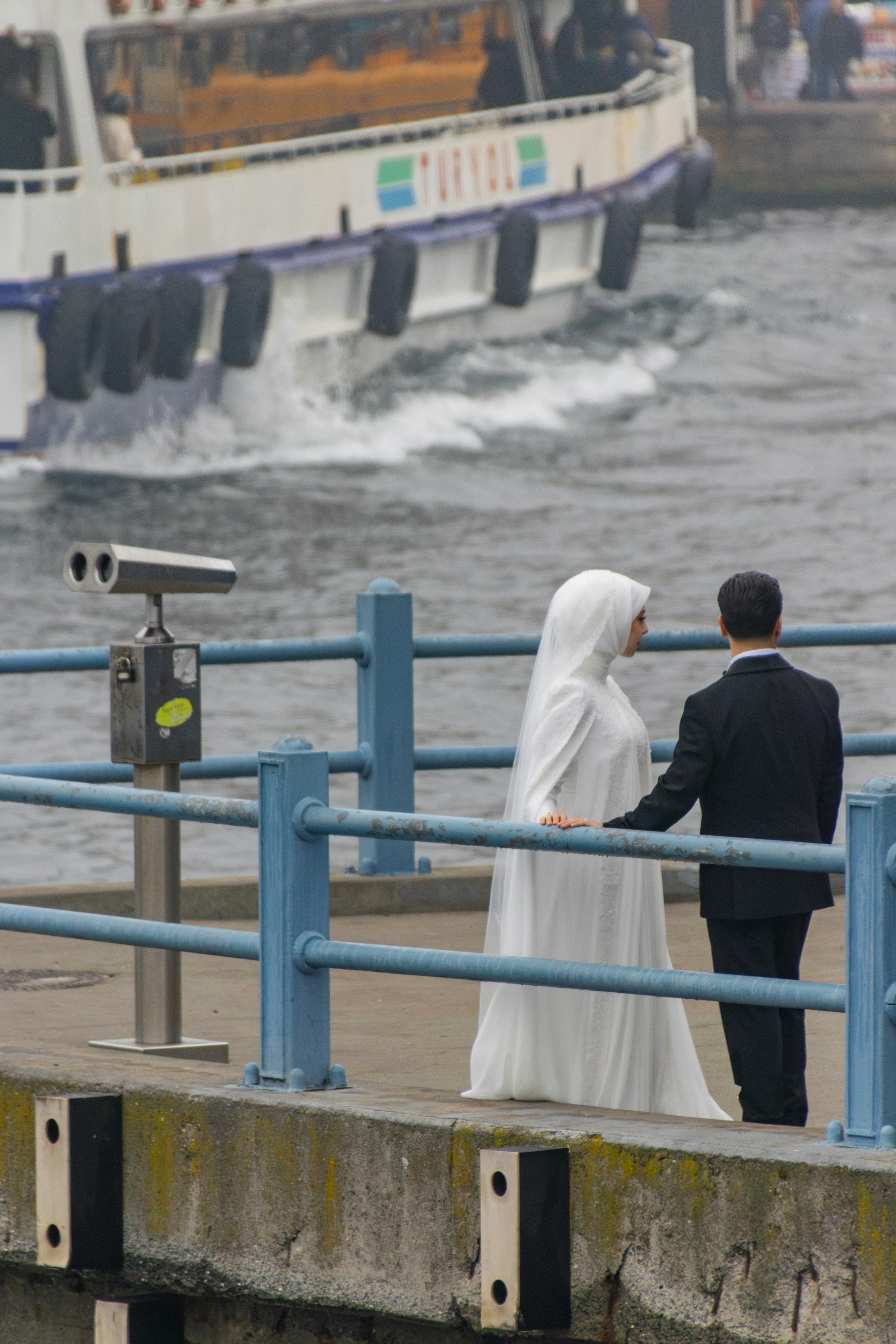 Bride in a headscarf and groom looking at the sea from a pier with blue railings