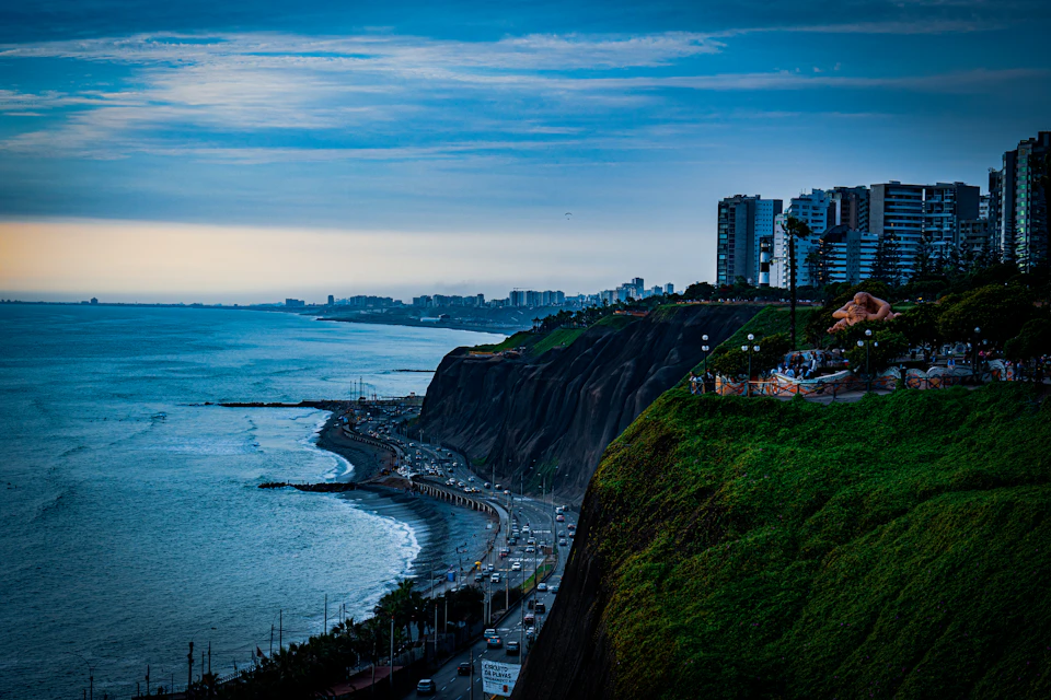 The image depicts a coastal cityscape with a cliff overlooking the ocean, featuring a mix of modern and traditional architecture. The city is situated on a hillside, providing a stunning view of the sea below. The sky above is painted in shades of blue, creating a serene backdrop for the urban setting. The perspective from the top of the cliff offers an expansive view of the city's skyline, with buildings of varying heights and designs scattered across the landscape. The image does not contain any discernible text or countable objects, but it captures the essence of a coastal city at dusk.