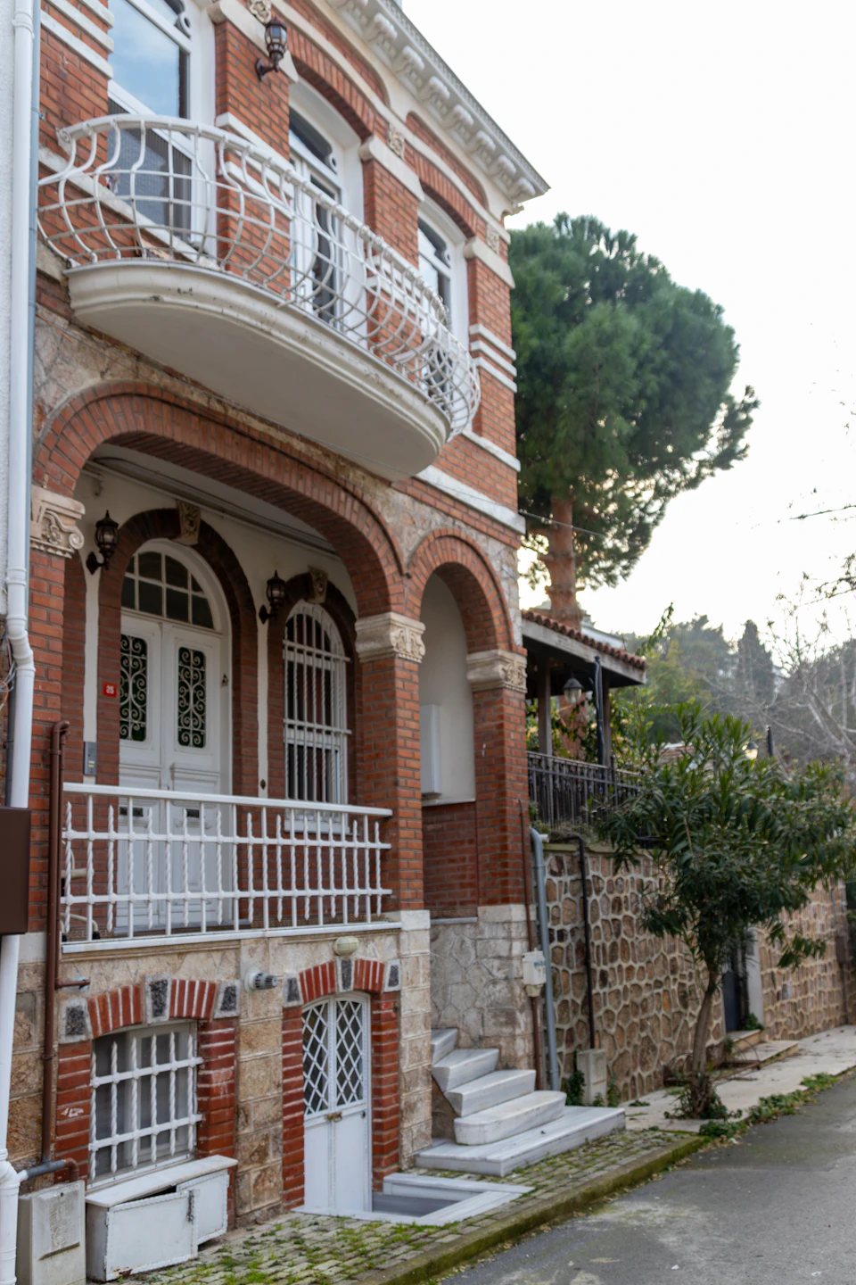 The image depicts a two-story brick building with a white balcony and arched windows, situated on a street corner. The building features a red brick facade that contrasts with its white trim. A tree is visible to the right side of the building, adding a touch of nature to the urban setting.

In front of the building, there are two sets of stairs leading up to the entrance, and a small garden can be seen on the ground floor. The sky above appears hazy, suggesting an overcast day.
