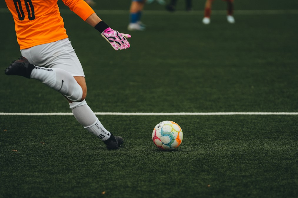 Un gardien en maillot orange et gants roses s’apprête à frapper un ballon multicolore sur un terrain synthétique