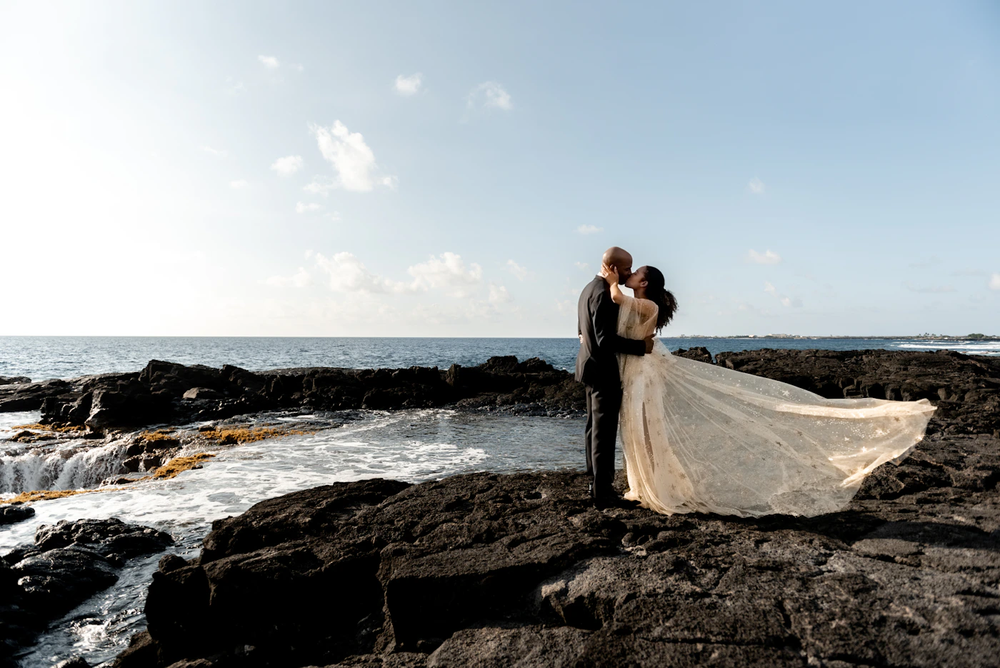Couple in elopement attire at the edge of a crystal-clear tropical beach with white sand and sea life visible in the shallow turquoise water
