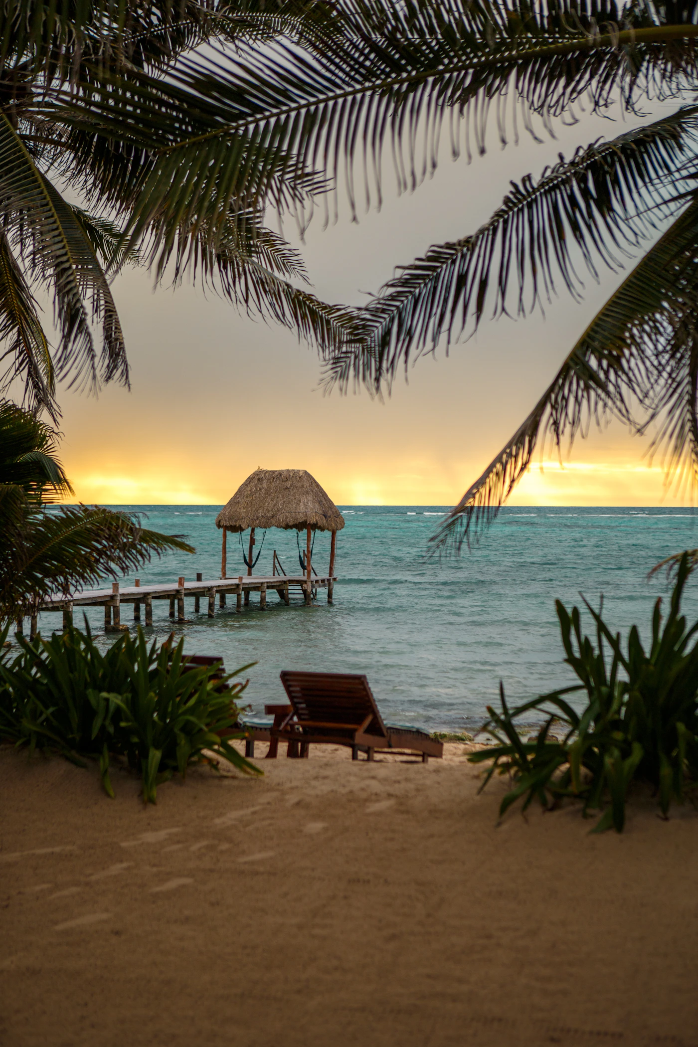 A palapa-roofed dock extending over turquoise Caribbean water at golden dusk, framed by palm fronds above, warm golden-orange sky reflecting on the water