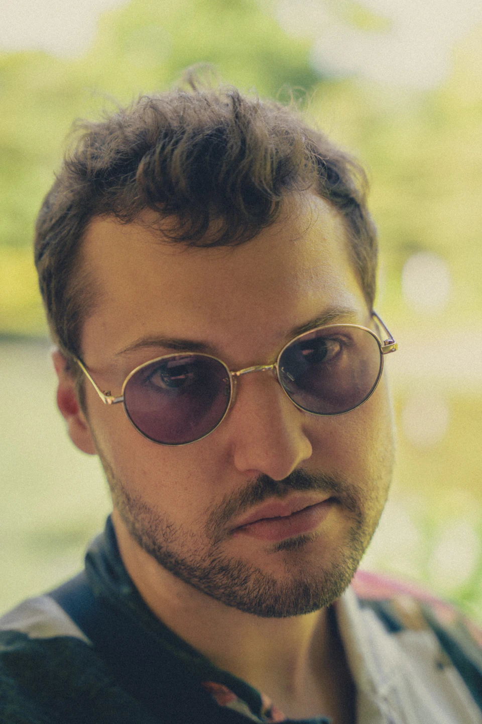 The image shows a man wearing round sunglasses and a black jacket, with his face partially obscured by the glasses. The background is blurred, suggesting an outdoor setting such as a park or garden.