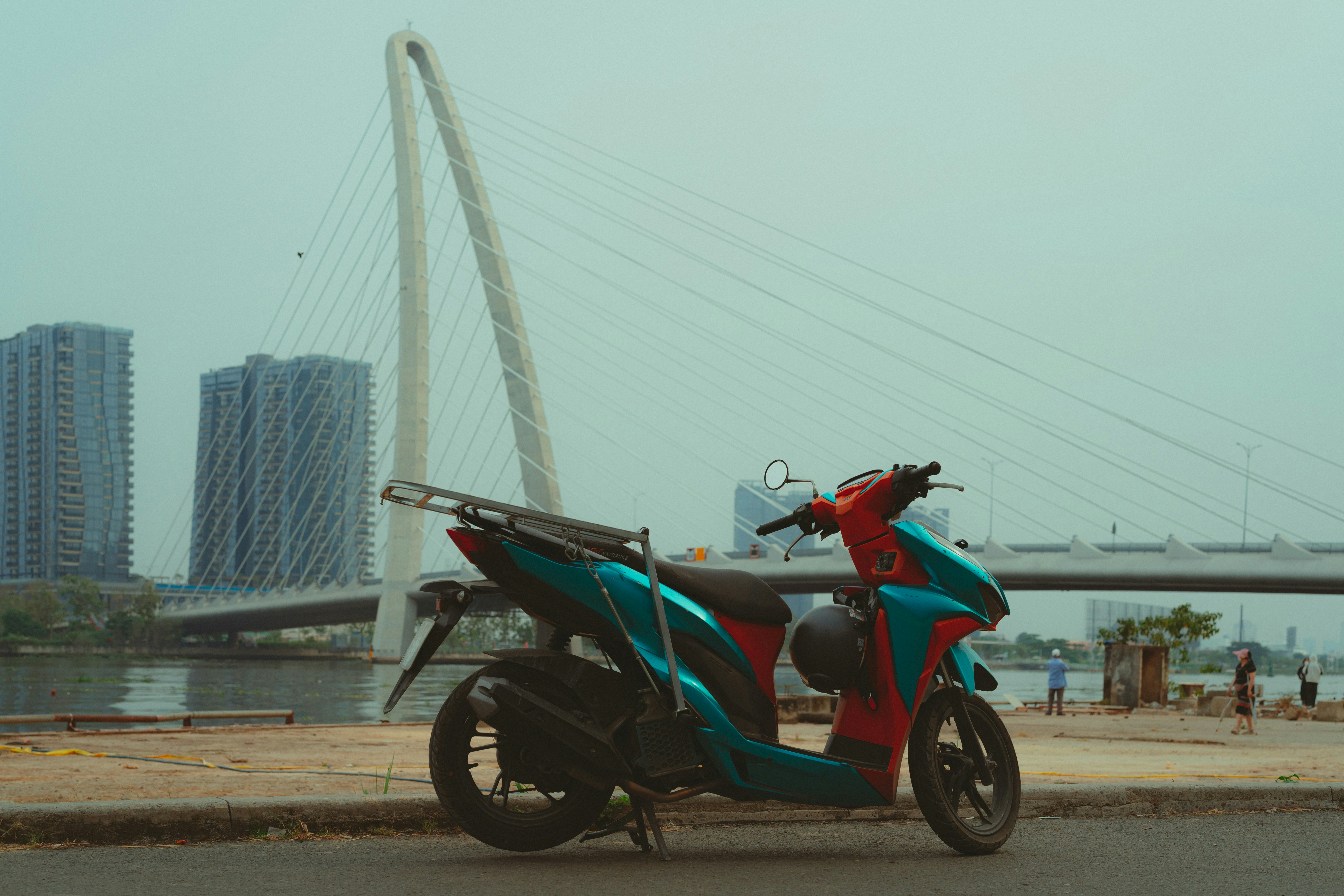 Skuter matik Honda biru-oranye dengan rak belakang terparkir di tepi sungai