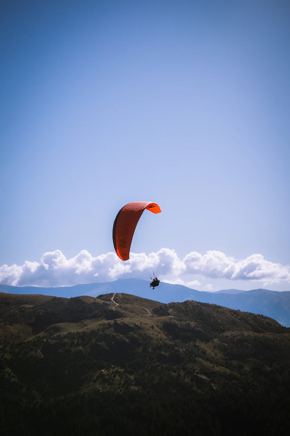 Red paraglider soars high above verdant mountain range under clear blue sky conditions