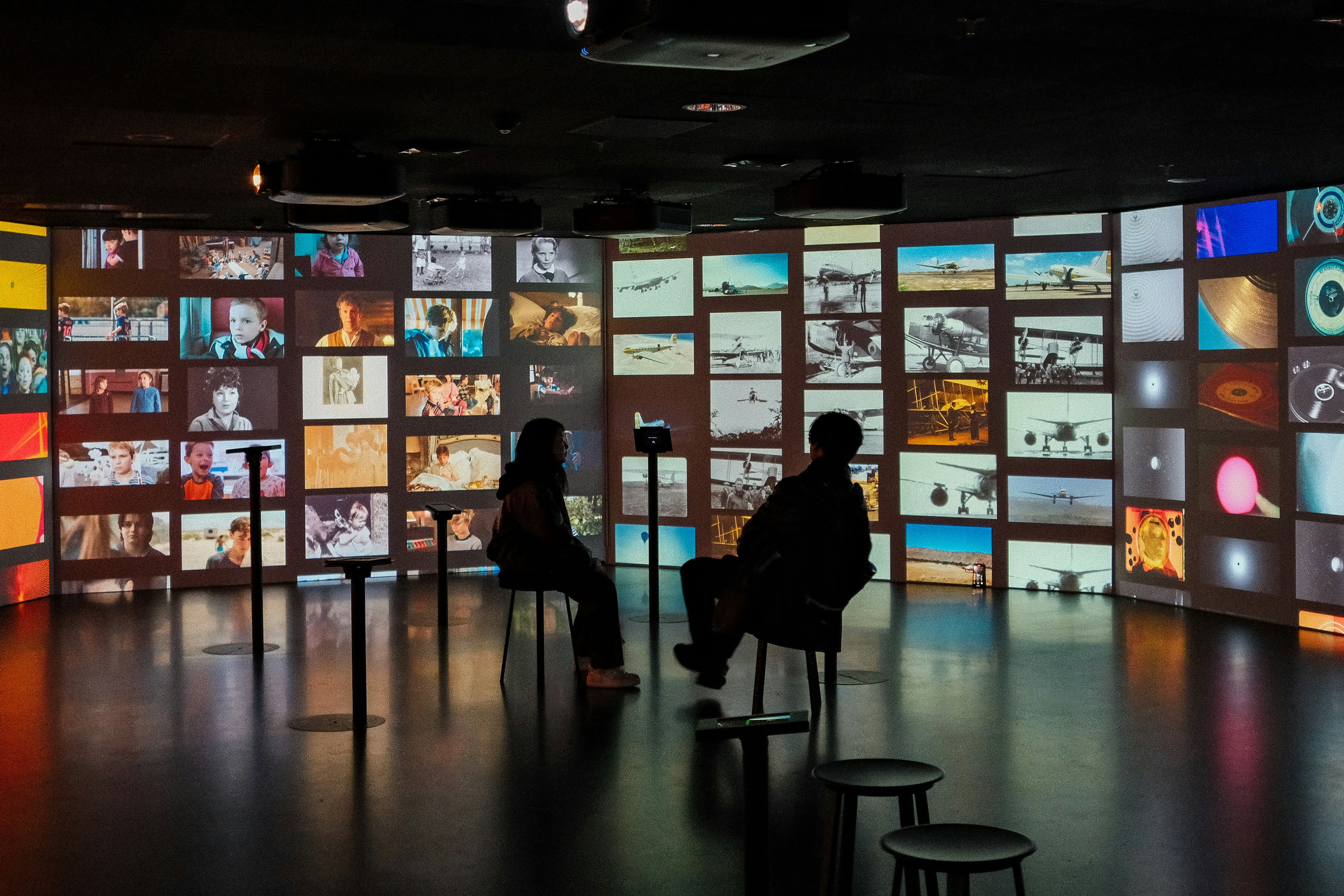 two people sitting in chairs in front of a wall of television screens