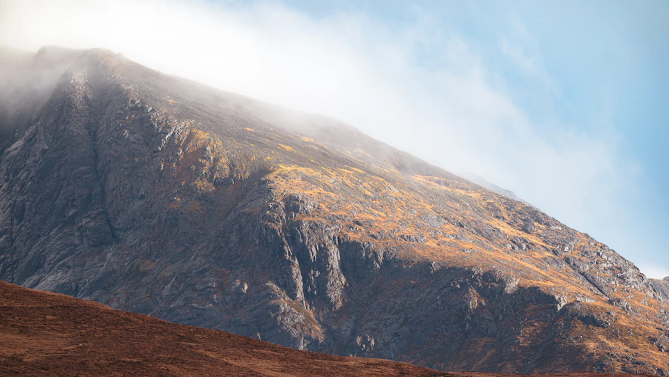 The image depicts a majestic mountain range with a rocky, gray cliff face rising into the sky. The mountains are covered in patches of green vegetation and brown rocks, adding depth to the scene. A light mist is present on the left side of the image, creating an ethereal atmosphere over the landscape.