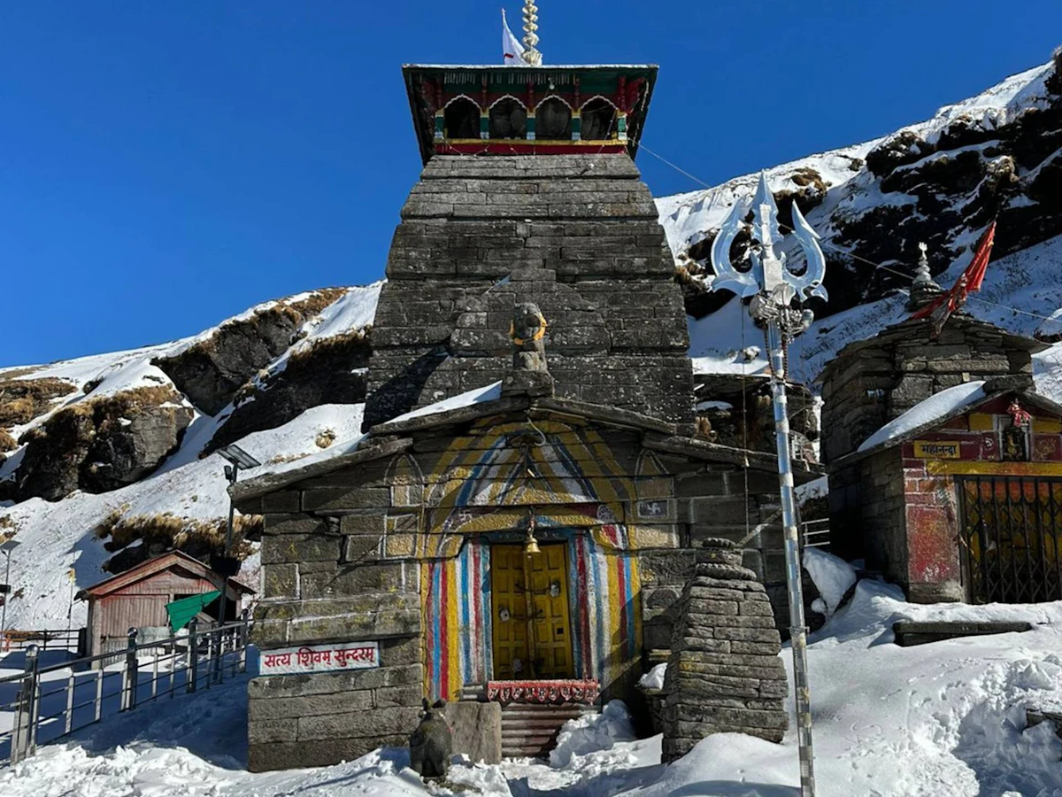 A stone Shiva temple with a painted doorway and a large trident outside, surrounded by snow under a deep blue sky on the Chopta-Chandrashila trek in Uttarakhand