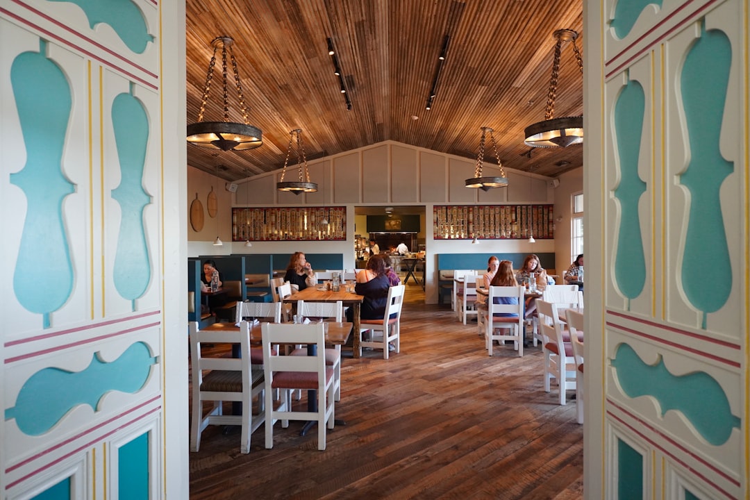 A view of a restaurant through an open door, A beautifully designed rustic restaurant interior with warm wood tones, handcrafted furniture, and inviting pendant lighting. The open dining space creates a cozy and welcoming ambiance, perfect for restaurant branding, hospitality marketing, and interior design inspiration. Ideal for showcasing modern farmhouse aesthetics, boutique dining experiences, and social gatherings.