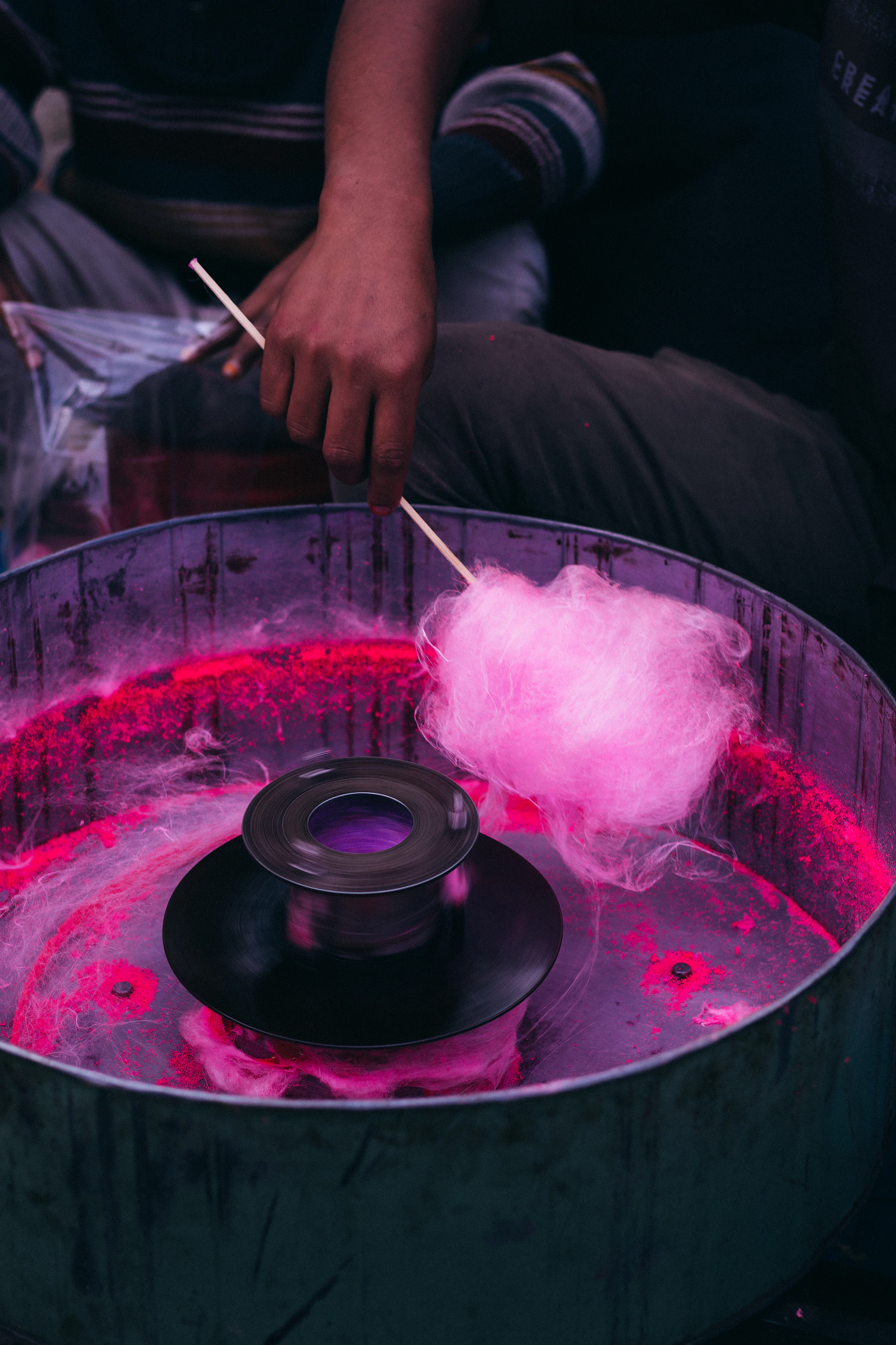A hand twirling a stick through pink floss as it gathers in a metal machine bowl around the spinner head