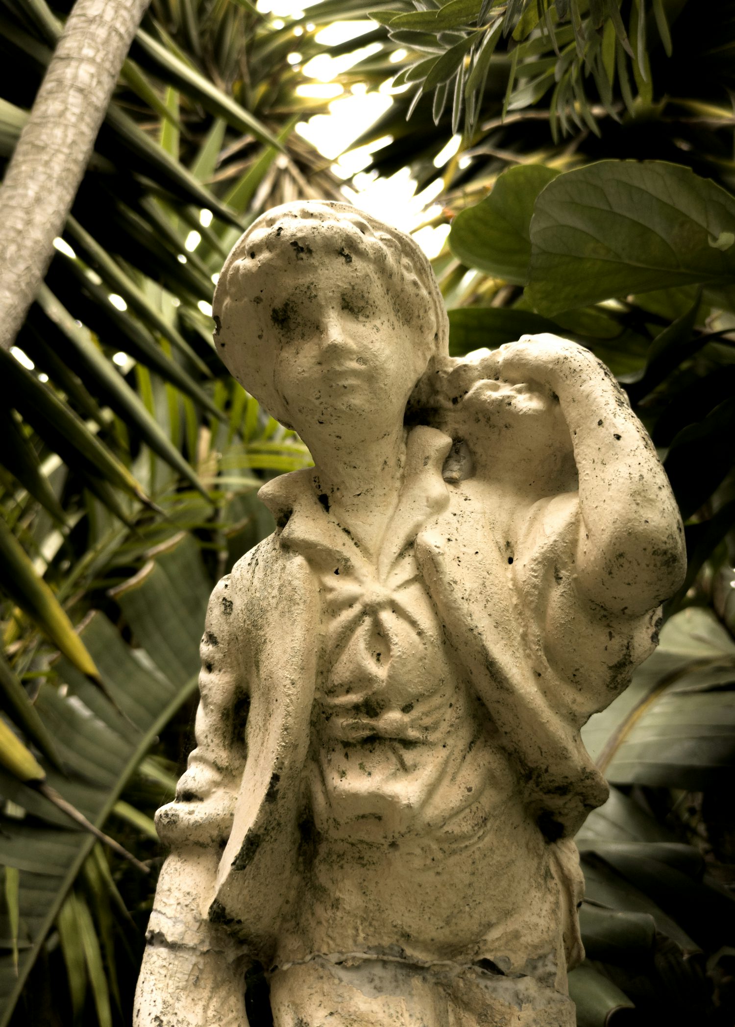 A close-up of a white statue of a person carrying something over their shoulder. The statue is very pitted and weathered and behind it is the green foliage of a garden. The sky can be seen between the leaves.