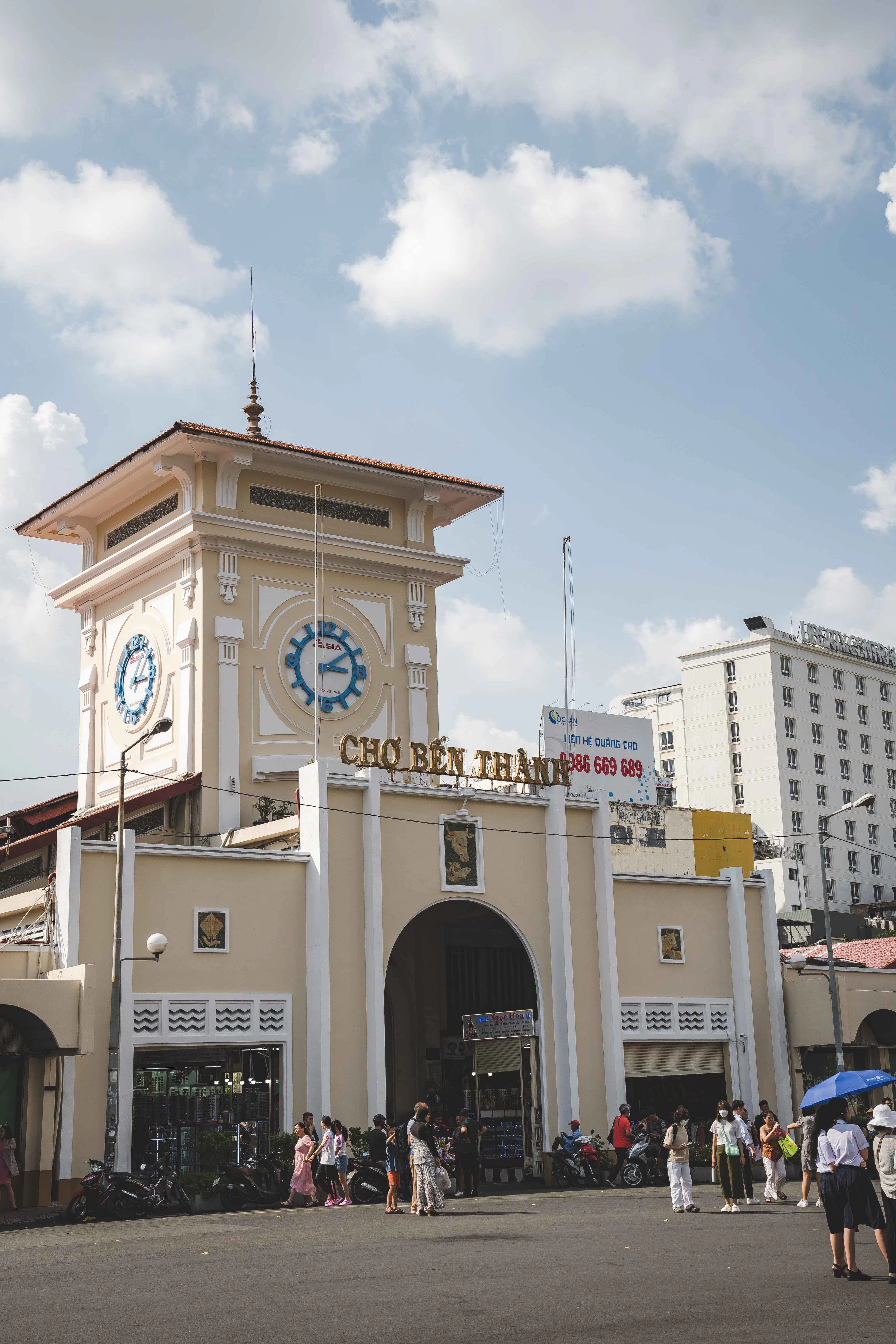 Ben Thanh Market's ochre clock-tower entrance under a bright blue sky, visitors gathered outside.