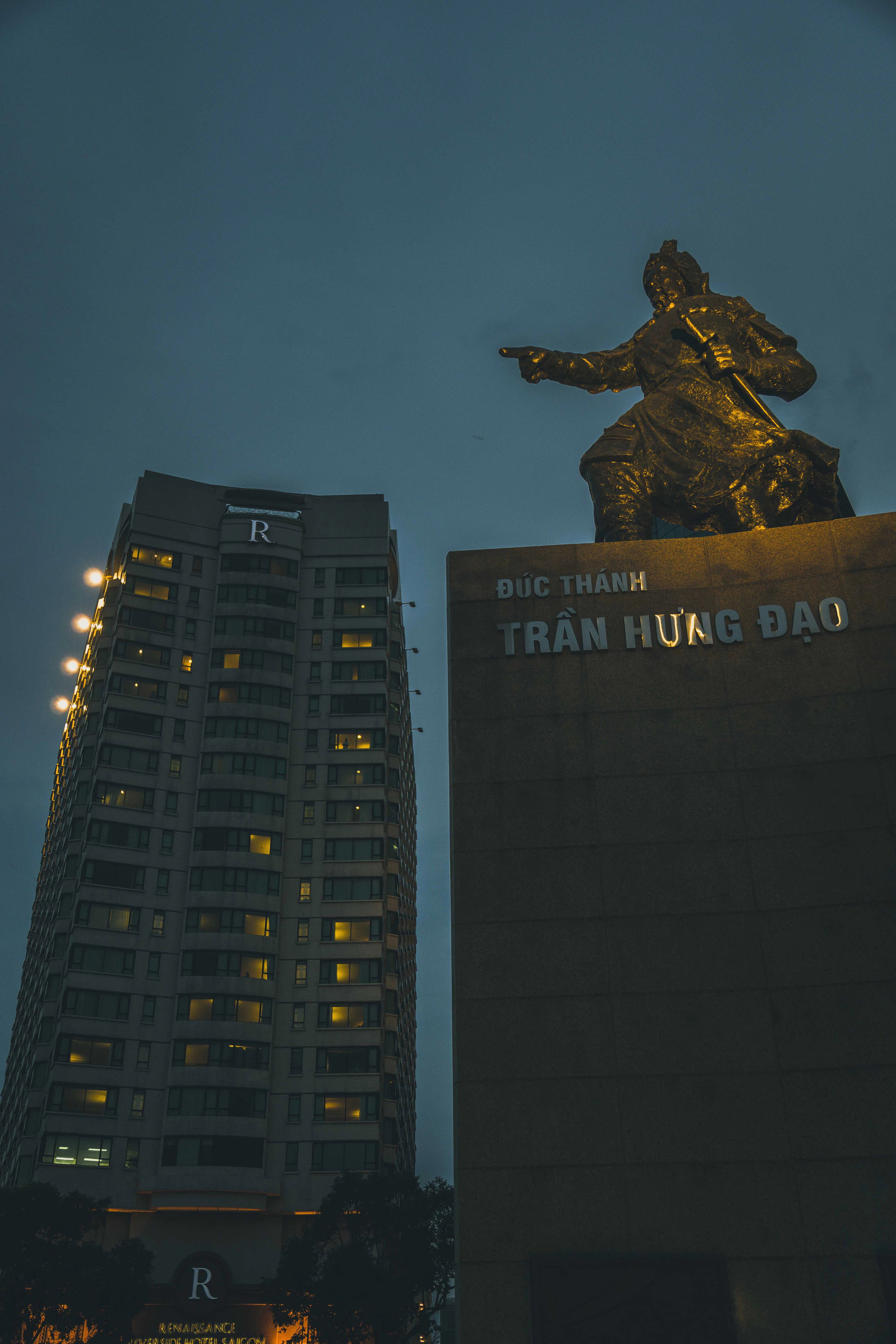 The gilded Tran Hung Dao statue stands against a dusk sky beside a lit riverside tower in Ho Chi Minh City.
