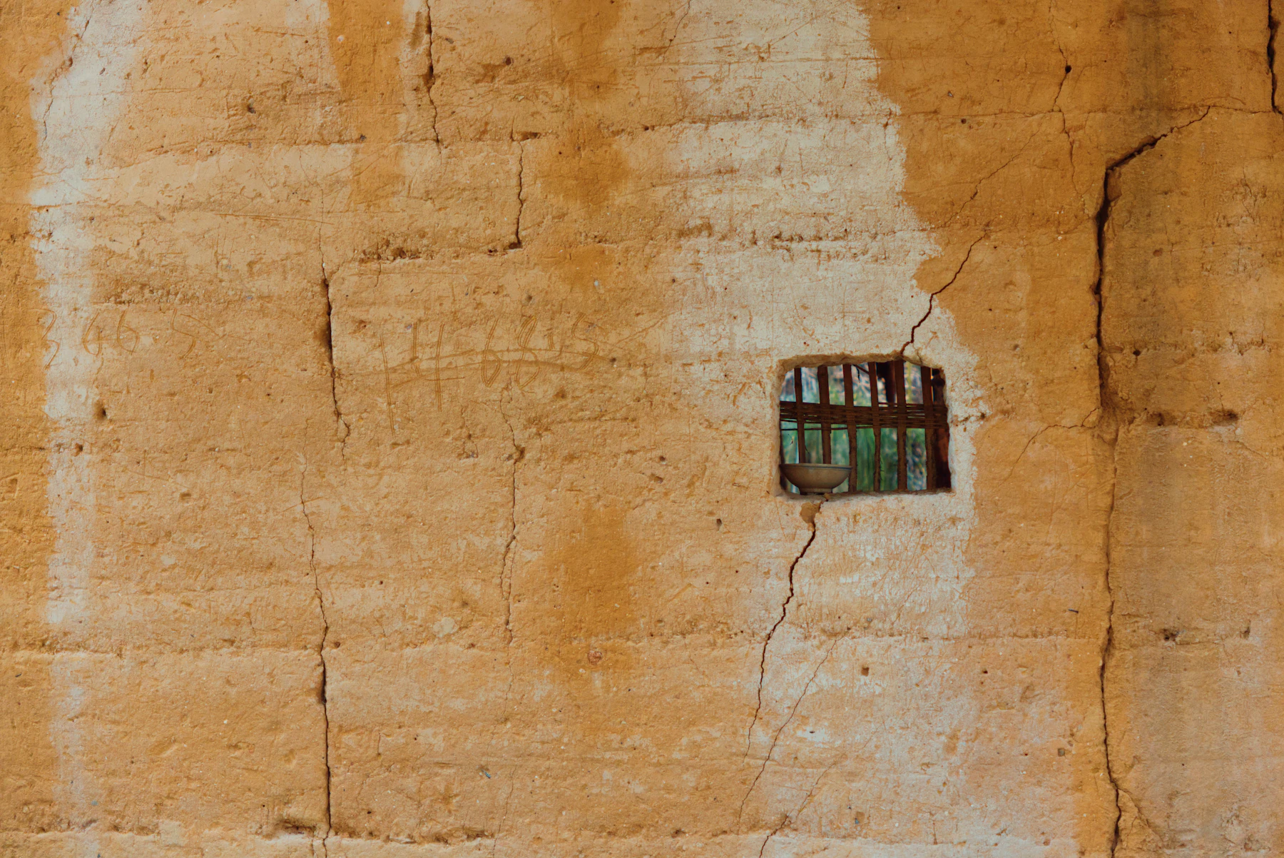 An ochre earthen wall with visible layer lines, hairline cracks and one small barred window
