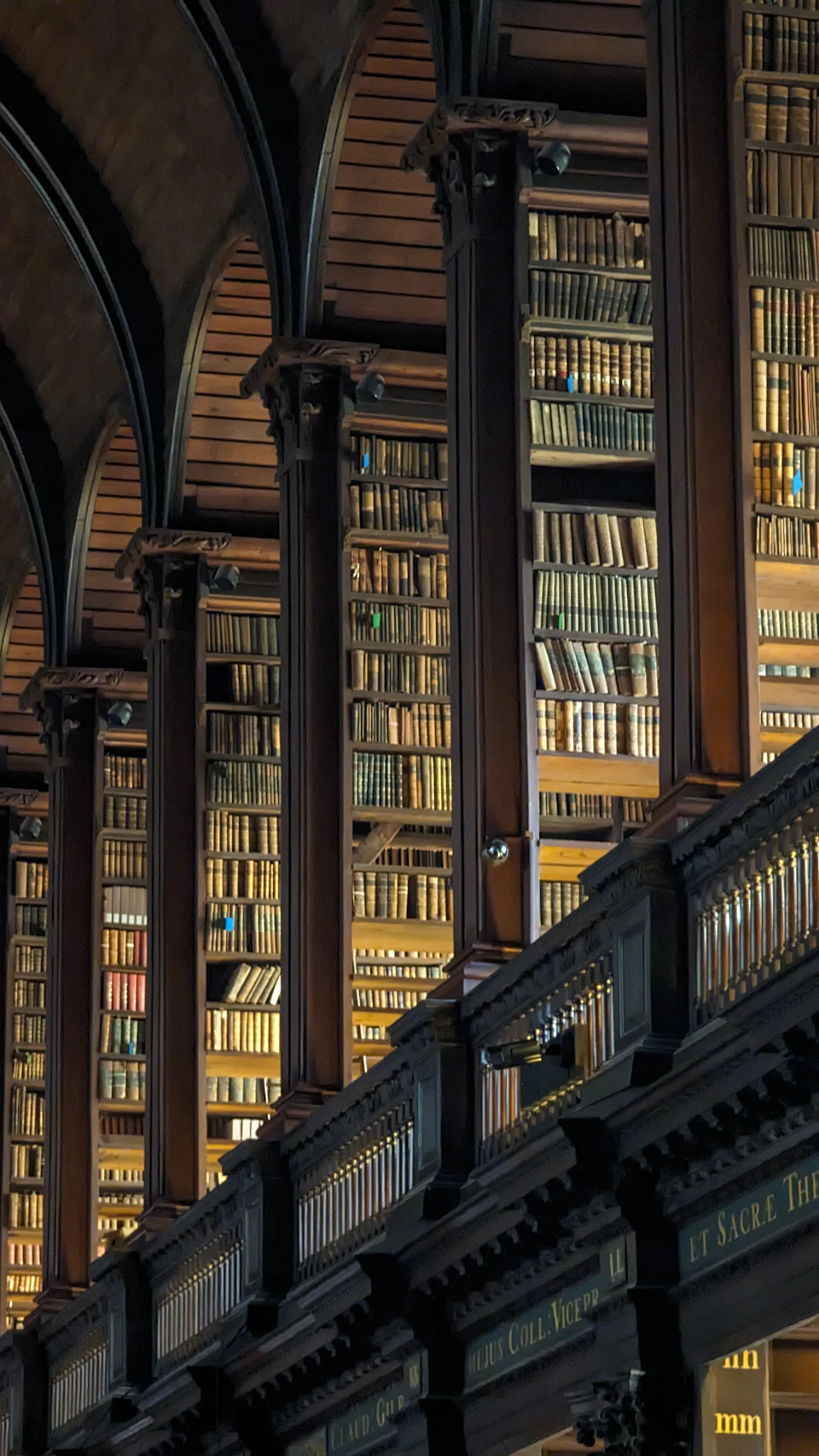 The image depicts a grand, old library with numerous tall bookshelves lining the walls and arched ceilings above them. The shelves are filled with rows upon rows of books, creating an atmosphere of knowledge and learning. The perspective is from below, looking up at the towering bookcases that stretch towards the ceiling. The lighting in the room is dim, casting a soft glow on the scene and highlighting the intricate details of the architecture and the books themselves.