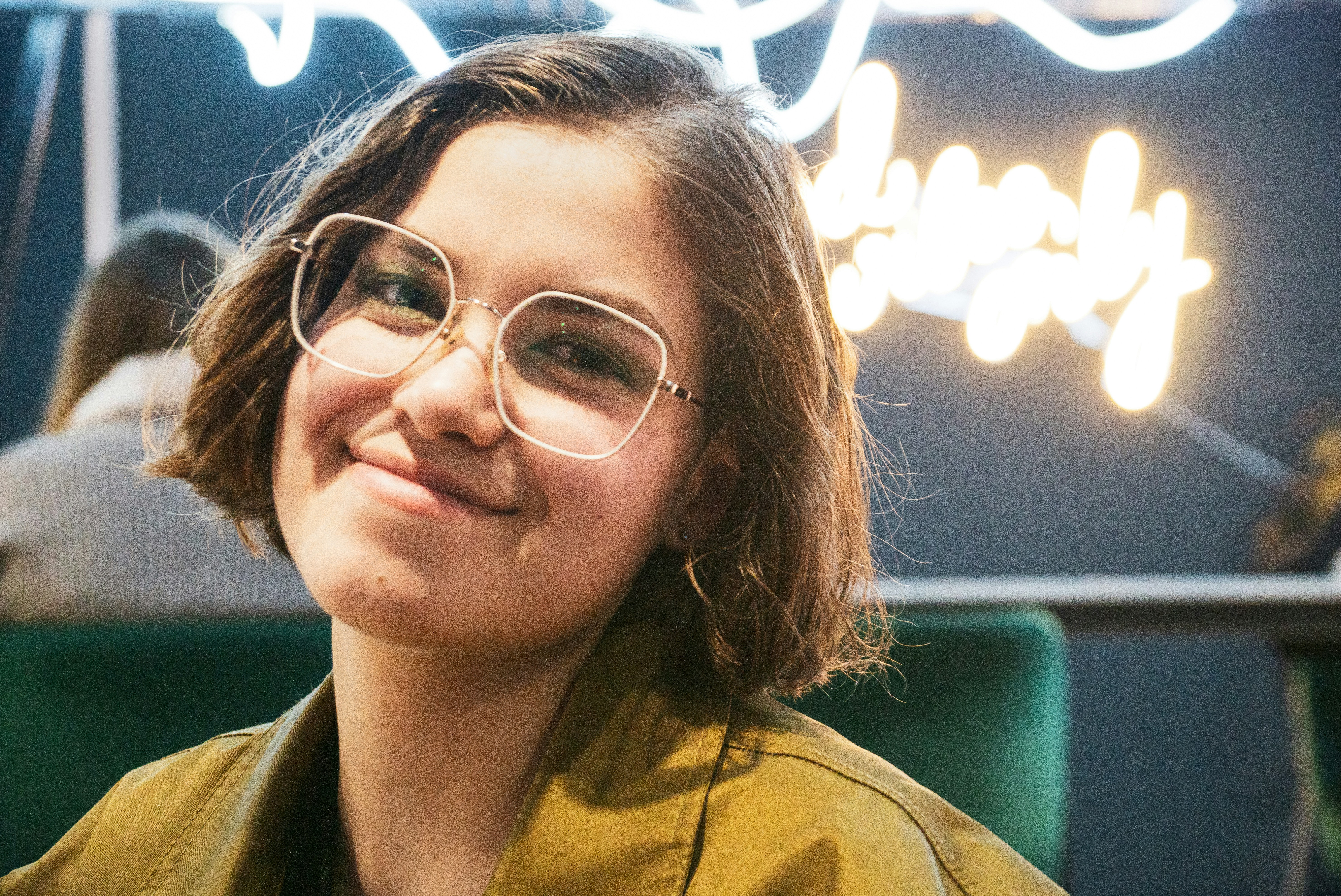 A girl with glasses smiles against a backdrop of decorative lights in a cafe.