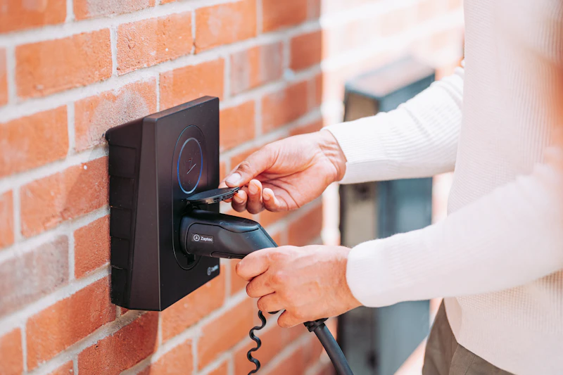 Hands plugging a charging cable into a black charge point mounted on a red-brick wall