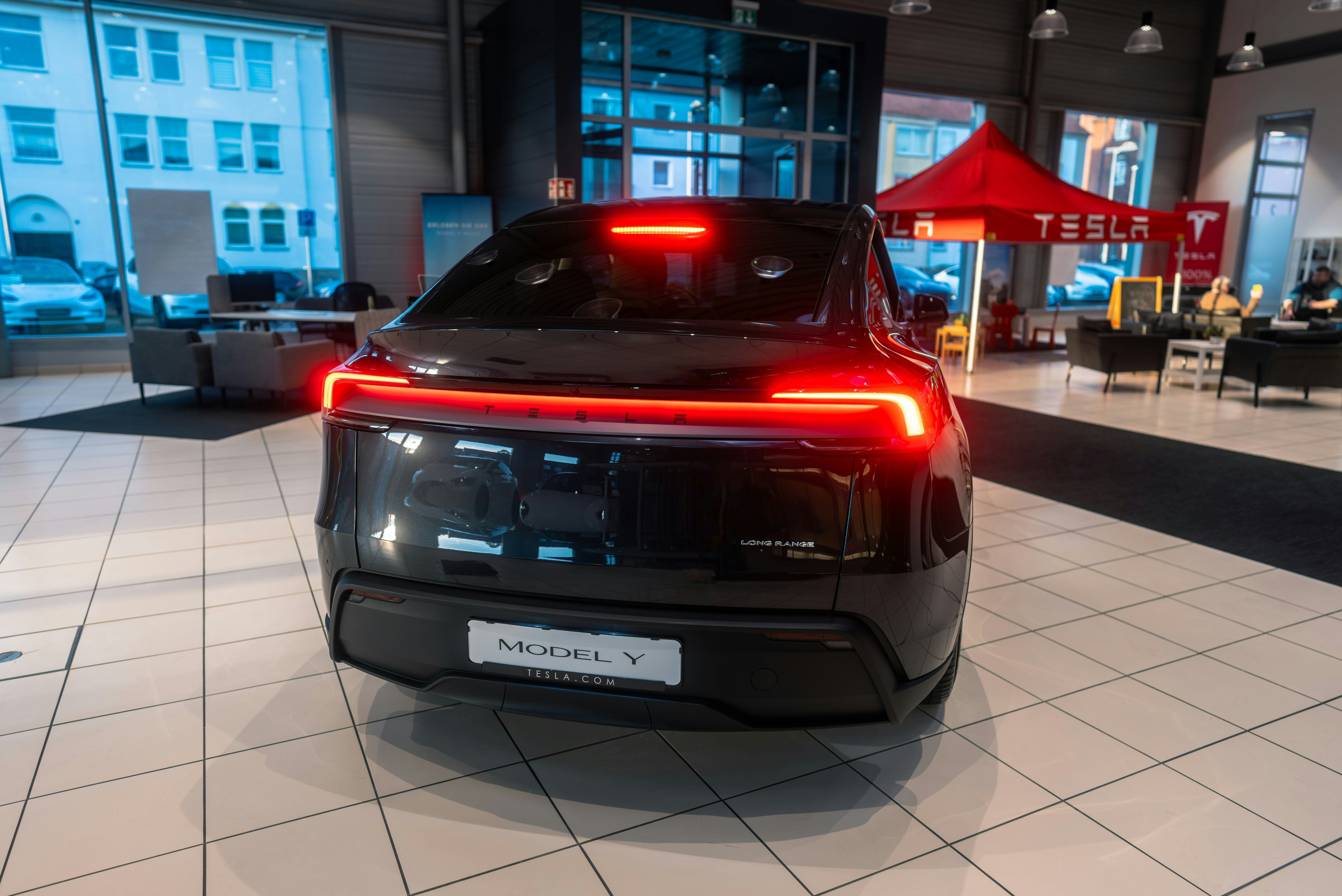 A person's hand holding a smartphone displaying the Tesla app, with a blurred image of a Tesla car in the background.