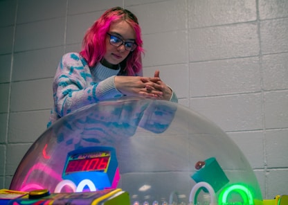 A woman with pink hair and glasses standing in front of a large object