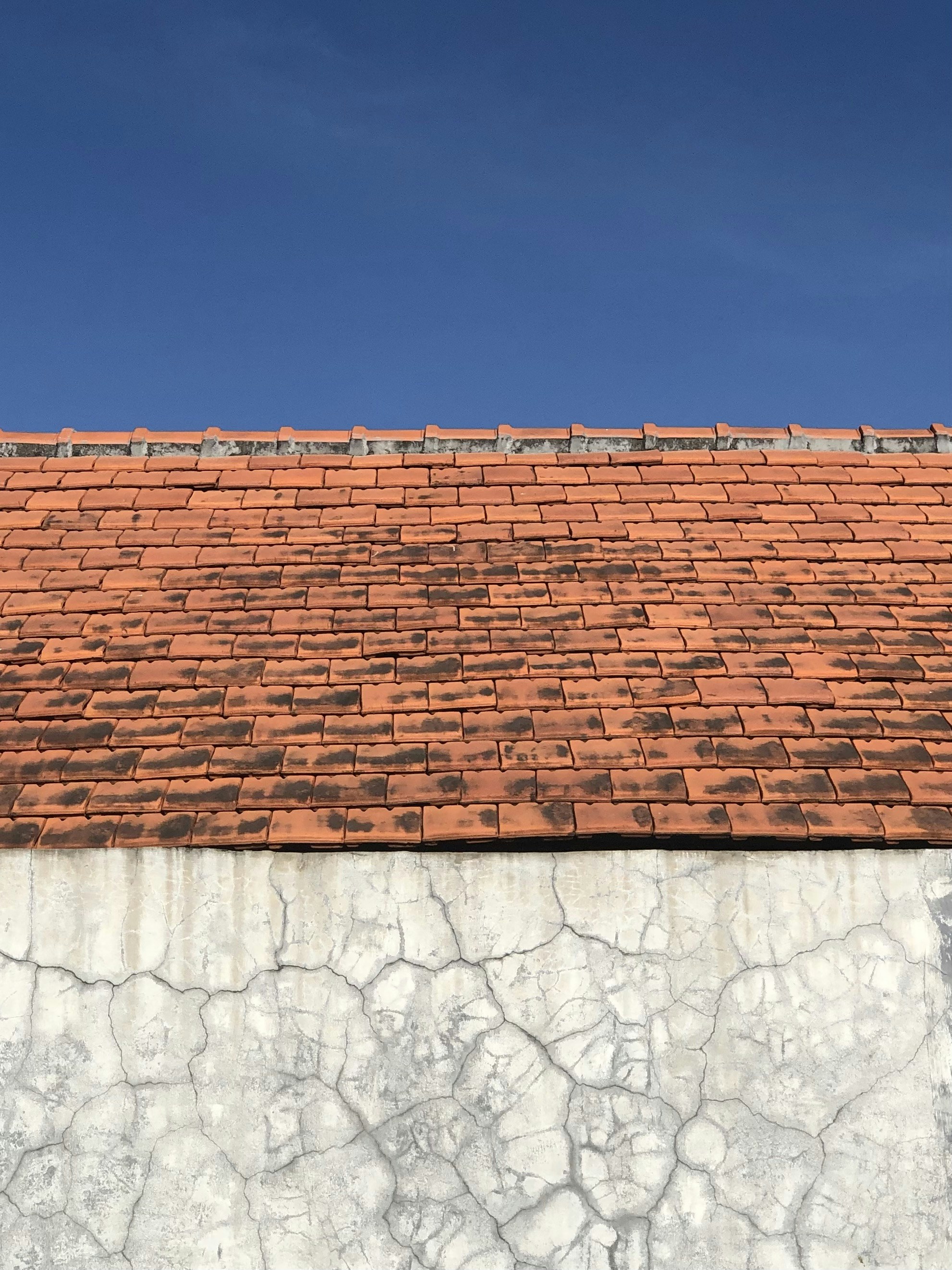 This photo showcases the upper portion of a weathered building against a clear blue sky. The roof is covered with aged, orange clay tiles, some of which appear darkened due to wear and exposure. Below the roof, the wall has a cracked and textured white surface, giving it an aged and rustic appearance. The contrast between the earthy tones of the roof, the rough texture of the wall, and the smooth blue sky creates a visually striking composition that highlights the beauty of imperfection and time-worn architecture.
