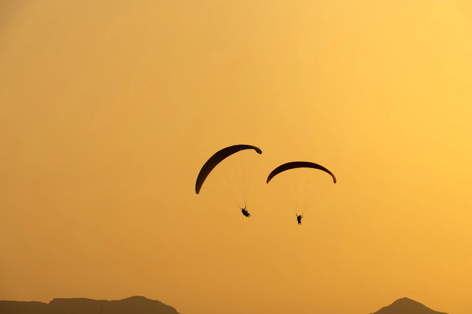 Two people paraglide in mid-air, angled towards each other, suspended above mountainous landscape