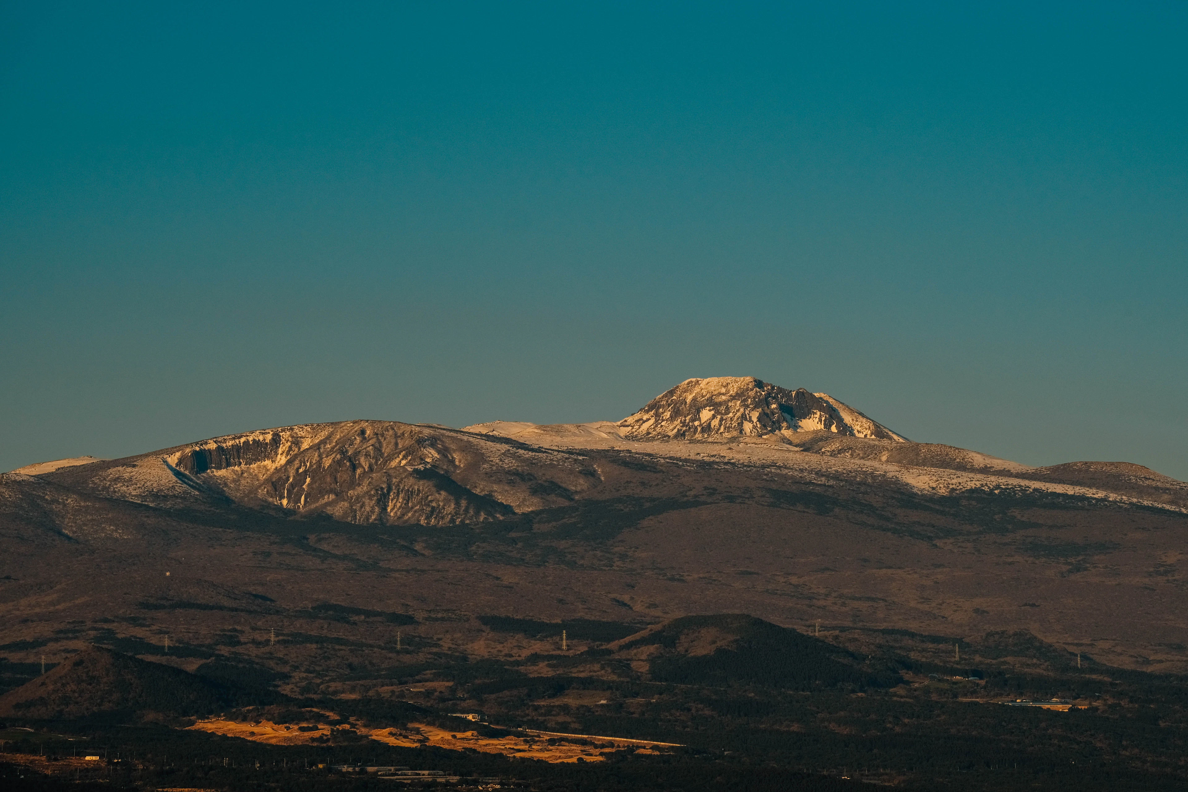 The snow-covered summit ridge and crater of Hallasan catching low golden light above the dark volcanic hills and farmland of southern Jeju Island, under a deep teal sky.