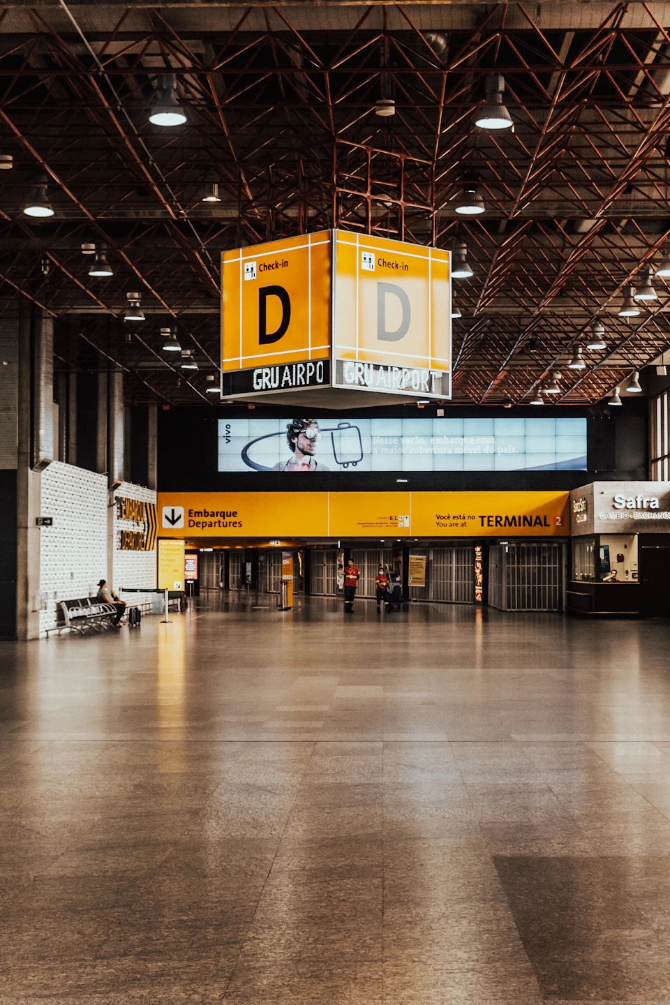The image depicts a large, empty airport terminal with a high ceiling and exposed beams. The terminal is illuminated by multiple lights that cast a warm glow throughout the space. A prominent yellow sign reading "D" stands out against the gray backdrop. In the foreground, there are two benches facing each other, one on the left side of the image and another on the right side.