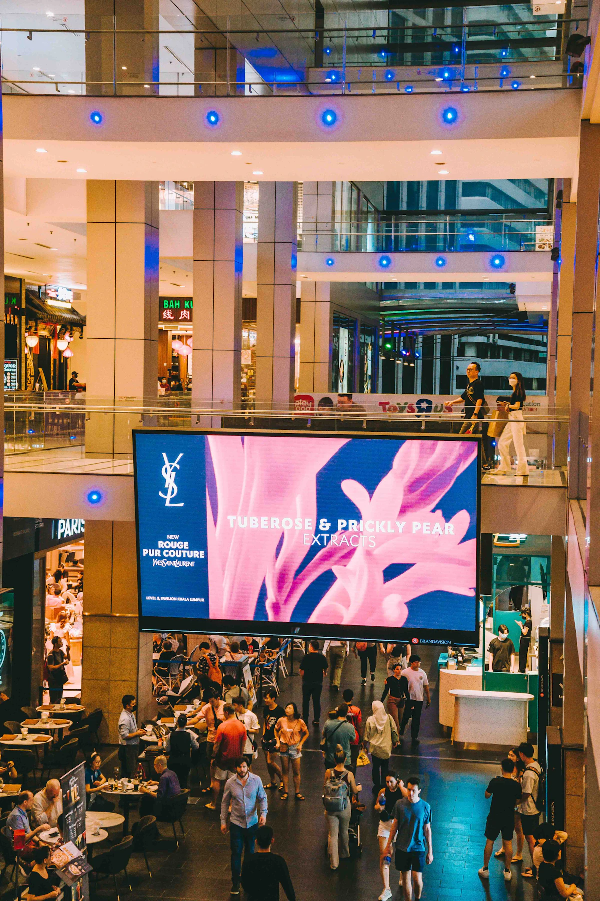 Pantalla publicitaria digital en el atrio de un centro comercial, con visitantes en las escaleras