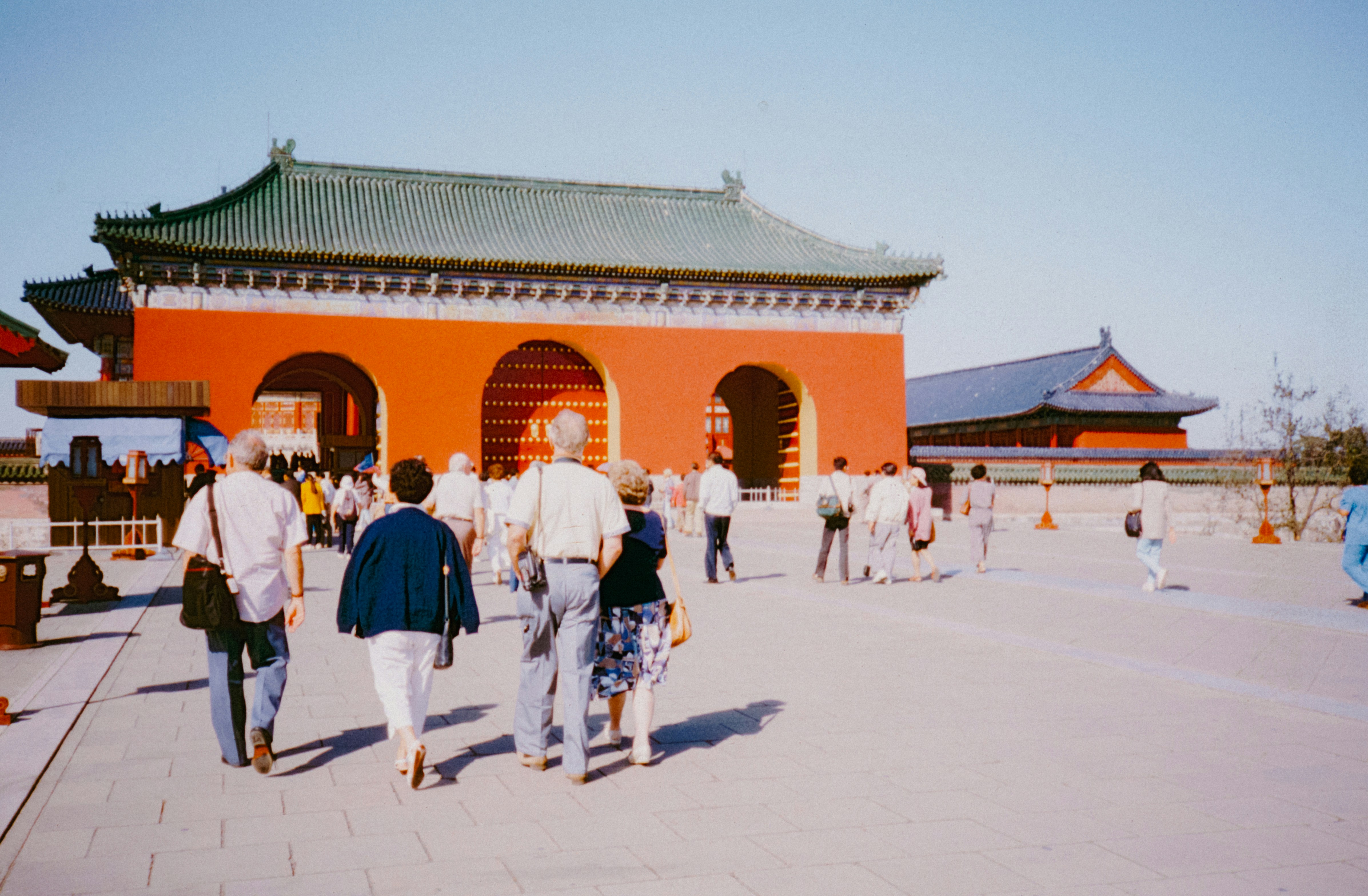 Tourists walk toward a traditional red Chinese gatehouse outdoors.